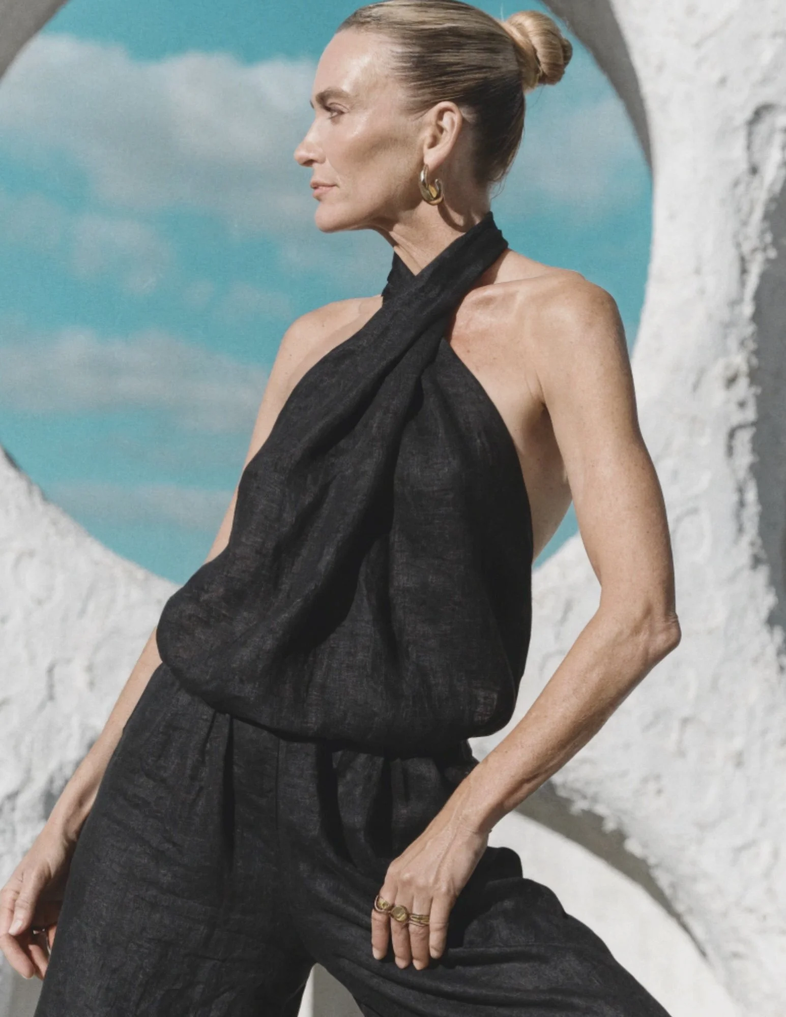 Photo of a middle age woman wearing a black linen halter pantsuit standing outside posing gracefully with a white brutalist sculpture behind her. The blue daylight sky and clouds peak through the sculpture's opening.