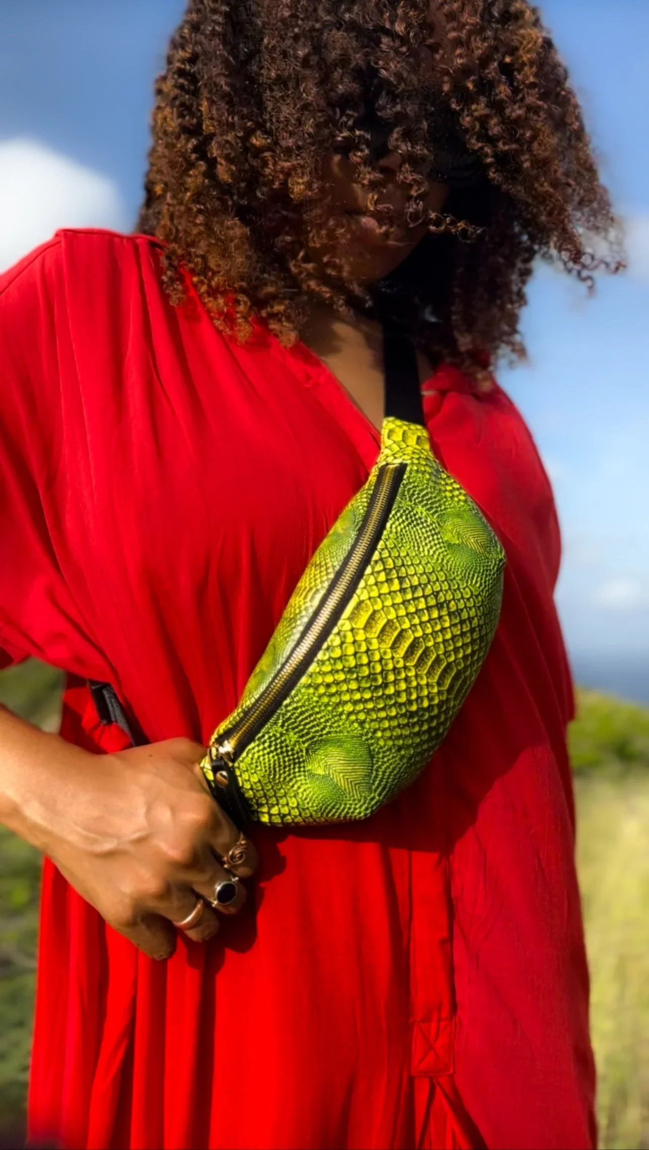 A close-up photo of a model with curly hair, wearing a red jumpsuit with a textured bright green Fanny Pack across her body, against a bright blue sky with white clouds.