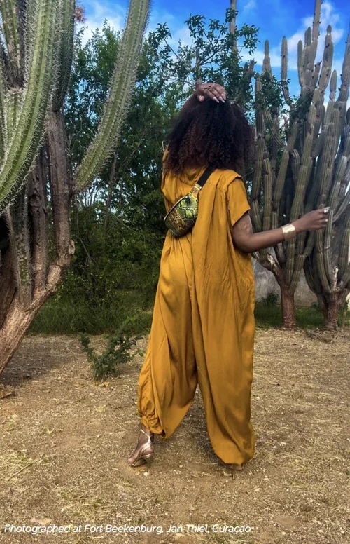 A photo of a model with curly hair wearing a yellow jumpsuit, and a green and black snakeskin print fanny pack across her body. The model is standing outside surrounded by green cacti and a blue sky with clouds.
