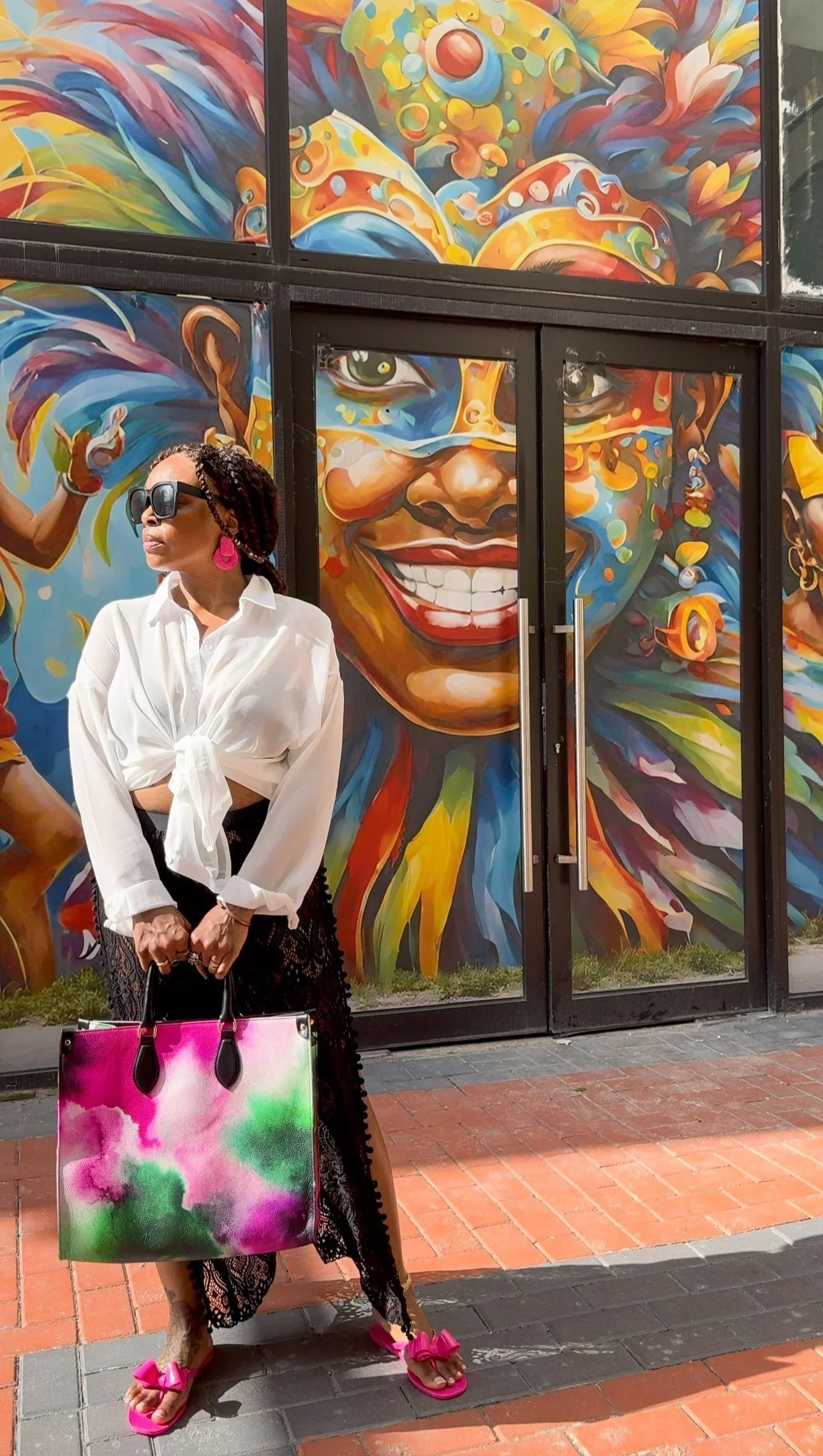 A photo of a model standing in front of a mural, wearing a white top and black lace pants, holding a pink, green, purple and white multicolored large leather shopper bag, with an image of a beautiful snake-haired woman on the side panel of the bag.