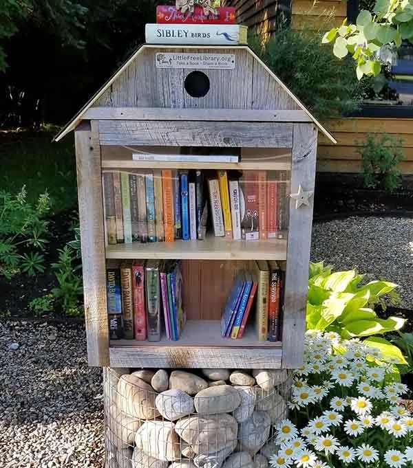 A small outdoor wooden library box filled with children's books, mounted on a stone base, surrounded by white daisies and green plants.