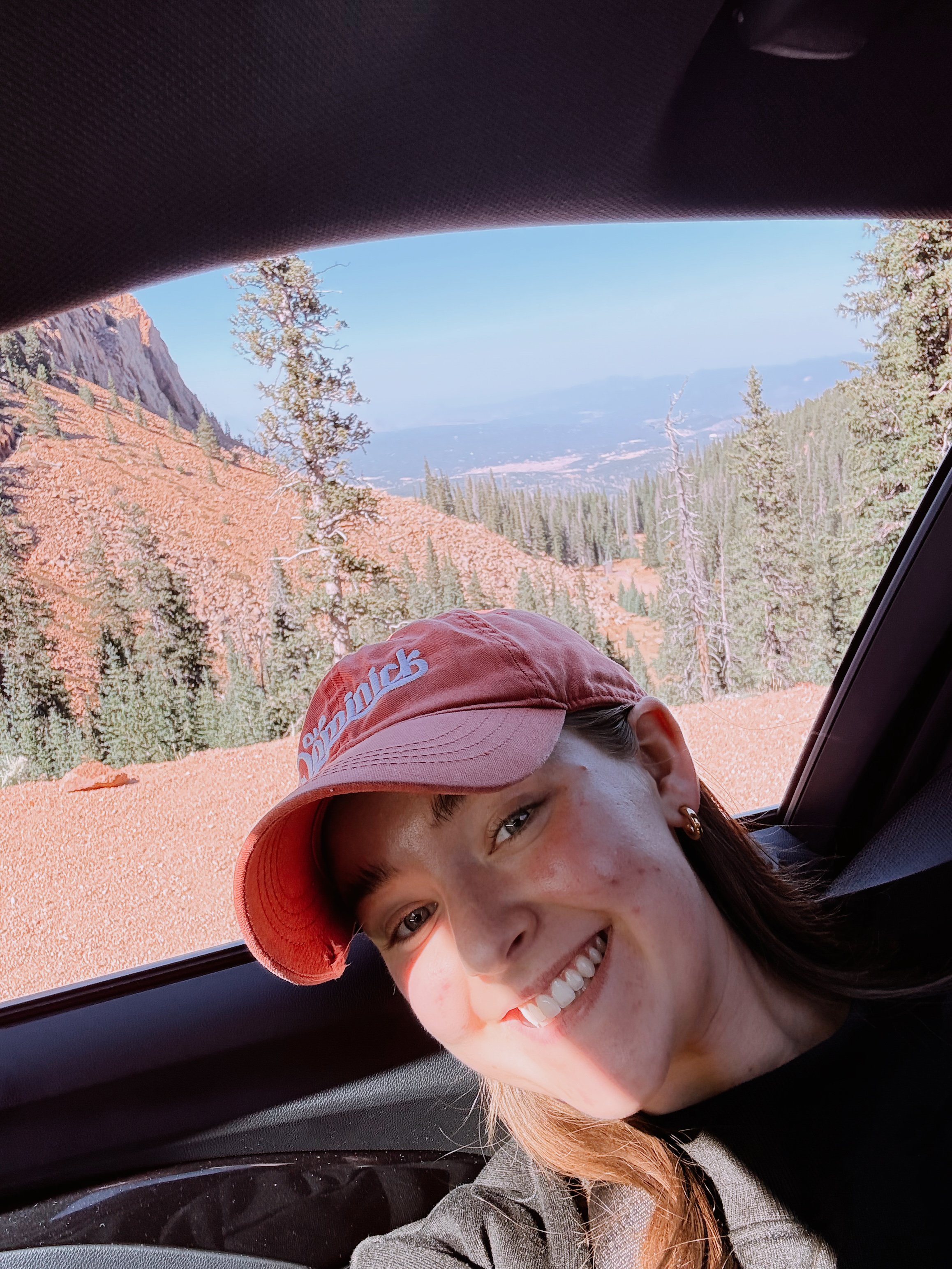A person wearing a red cap smiling inside a car with a mountainous view in the background.
