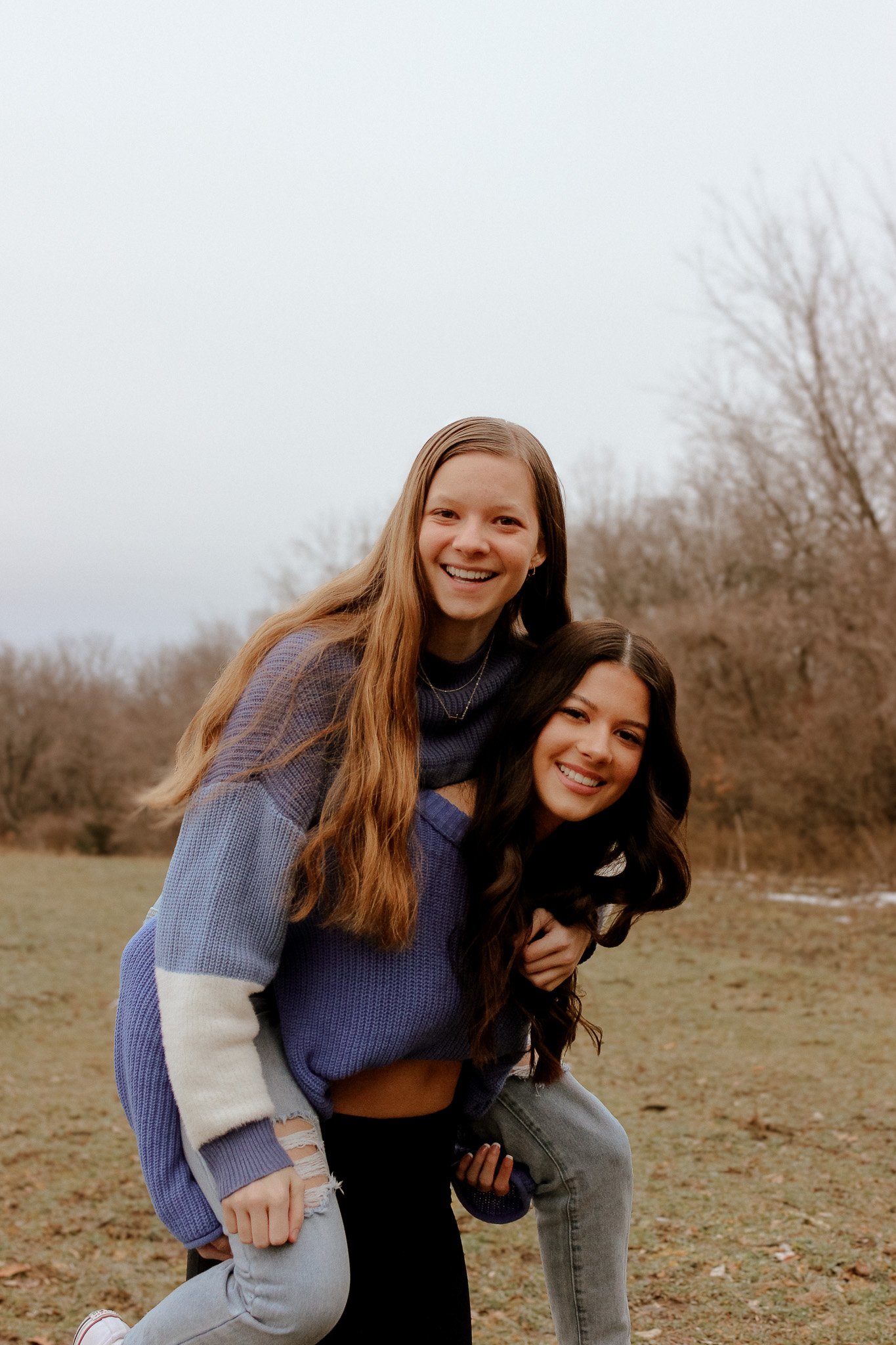 Two young women smiling, one giving the other a piggyback ride in an outdoor setting with bare trees and grass, wearing casual winter clothing.