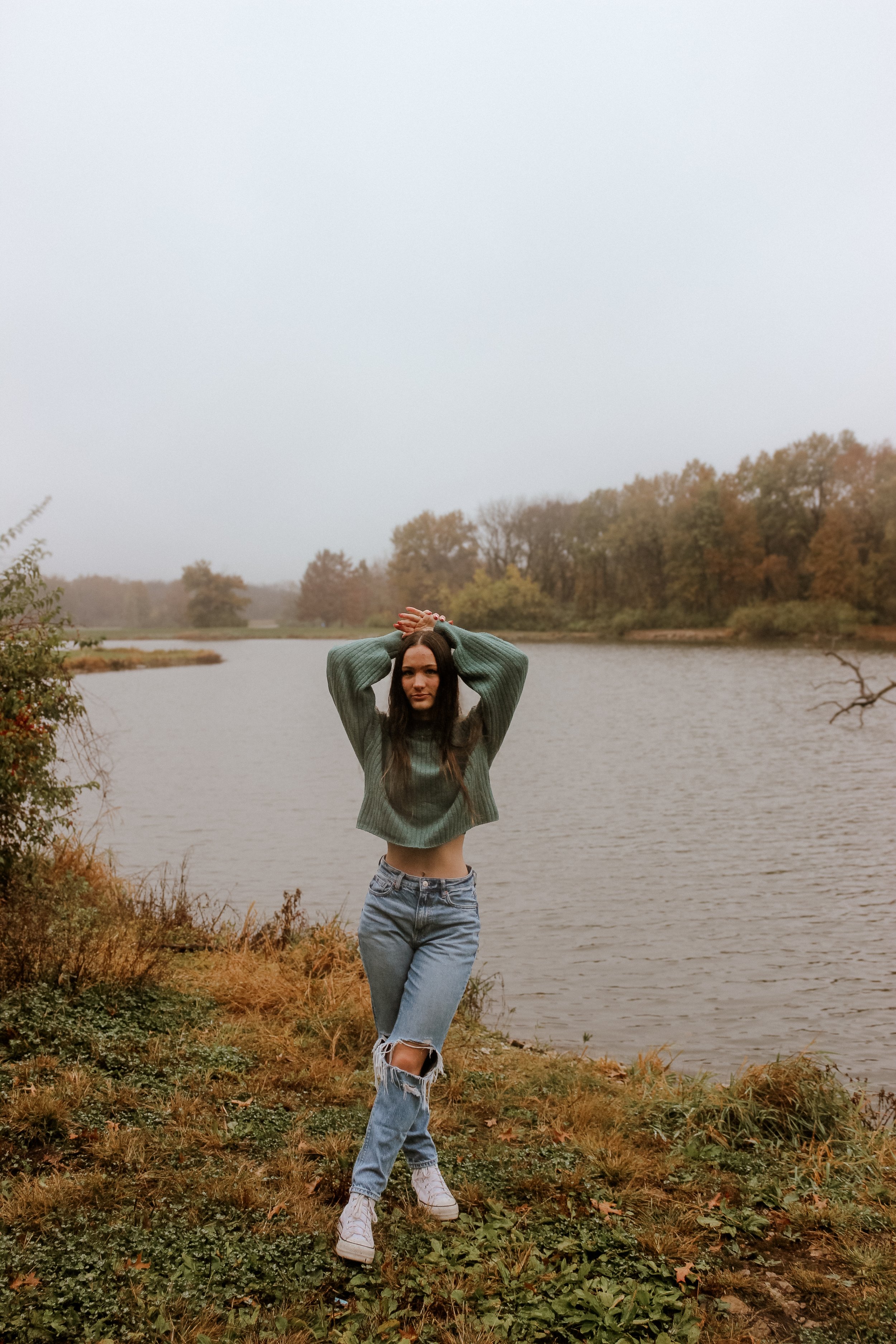 Person in a green sweater and ripped jeans standing by a lake, with arms raised over head, surrounded by autumnal foliage.