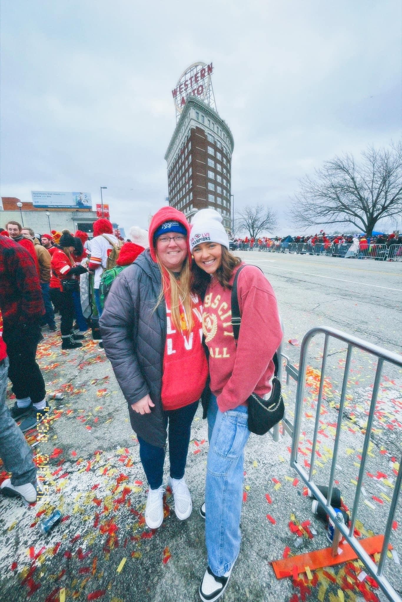 Two people at a parade with confetti on the ground, standing in front of the Western Auto building, wearing red sports apparel and winter hats.
