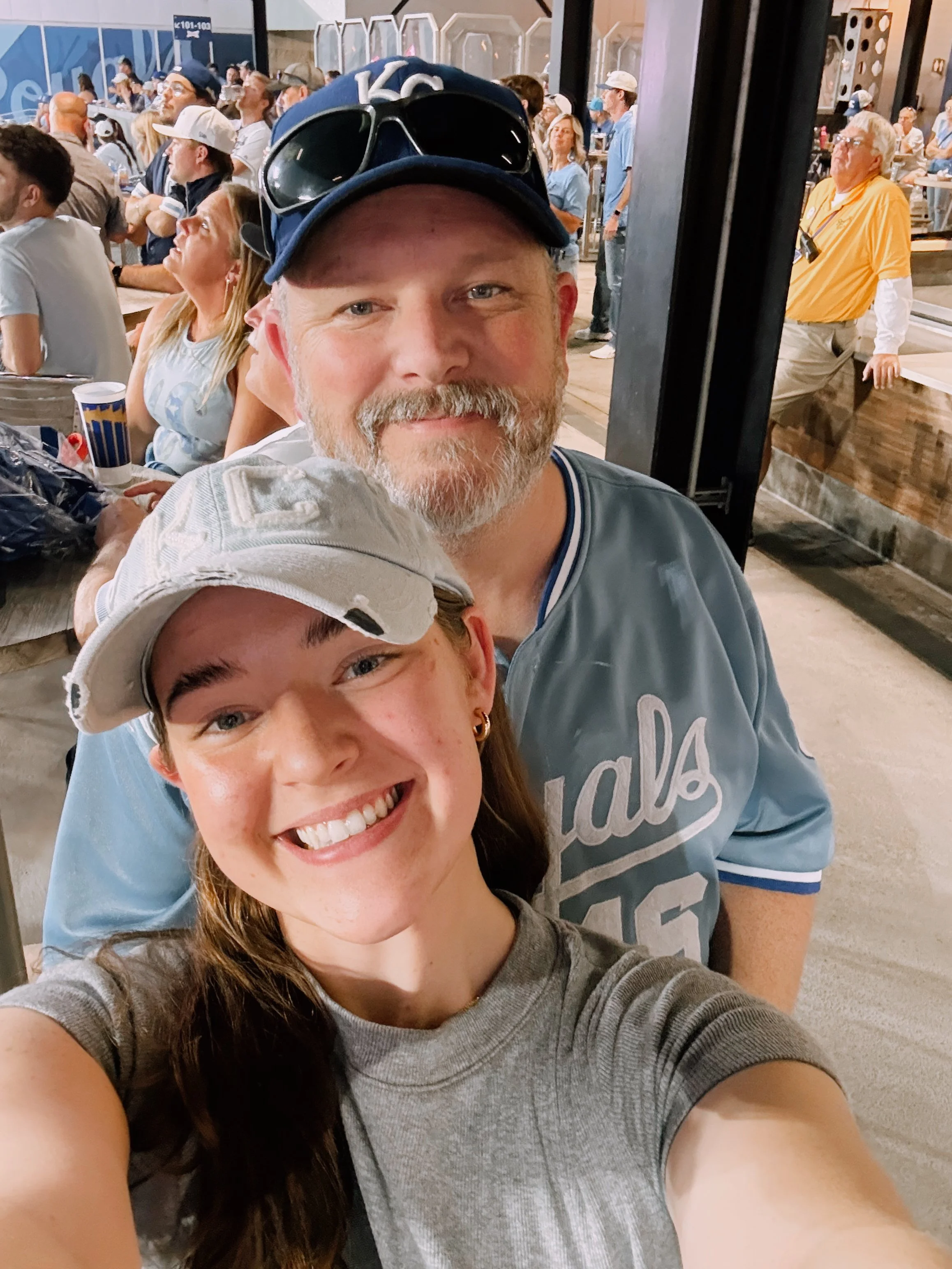 Two people smiling at a stadium, wearing baseball caps and Kansas City Royals shirts, surrounded by a crowd of spectators.