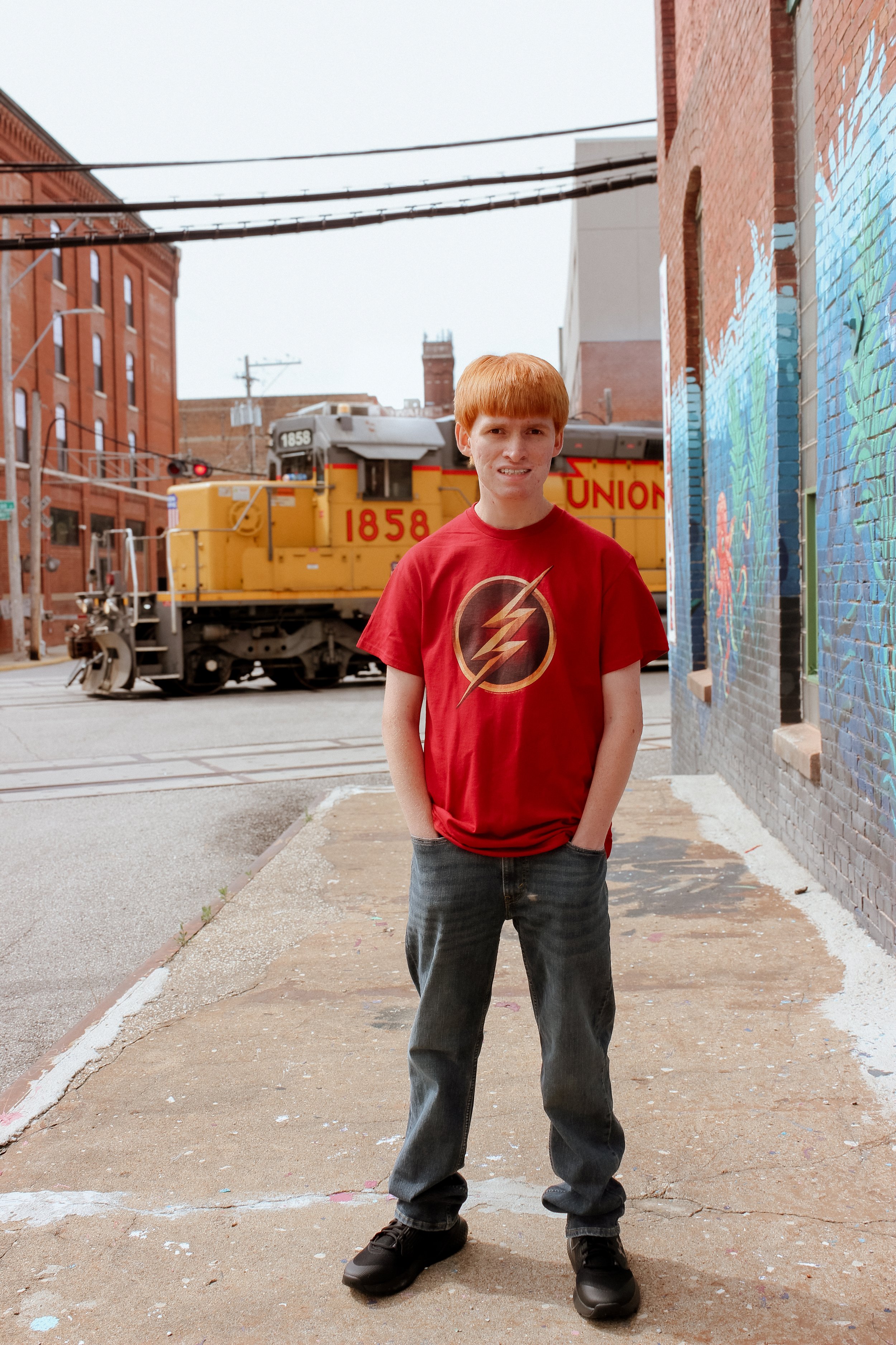 Person wearing a red Flash t-shirt standing on a sidewalk near a train, surrounded by urban buildings and colorful murals.