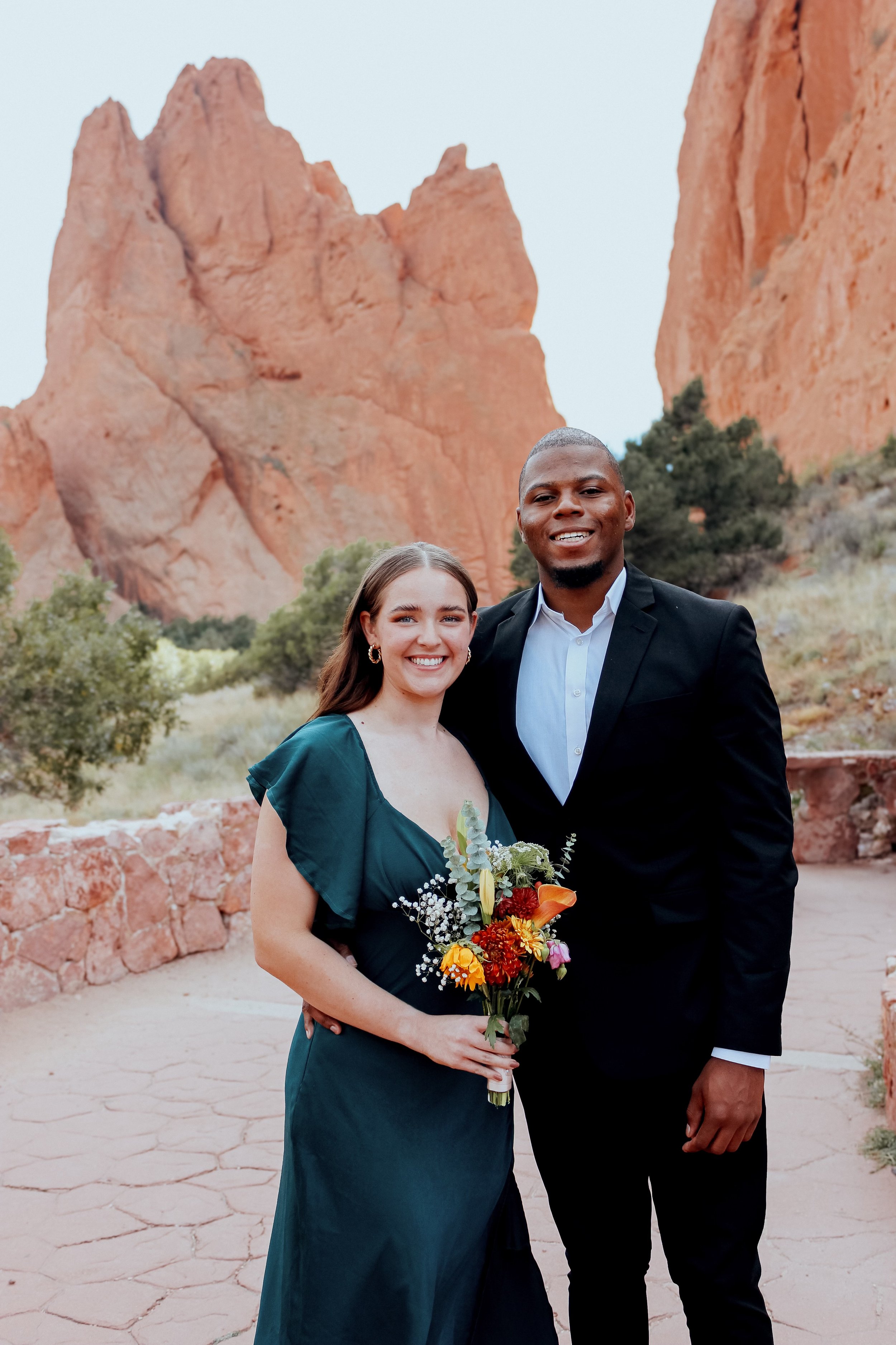 Couple posing outdoors, woman holding bouquet, red rock formations in background.