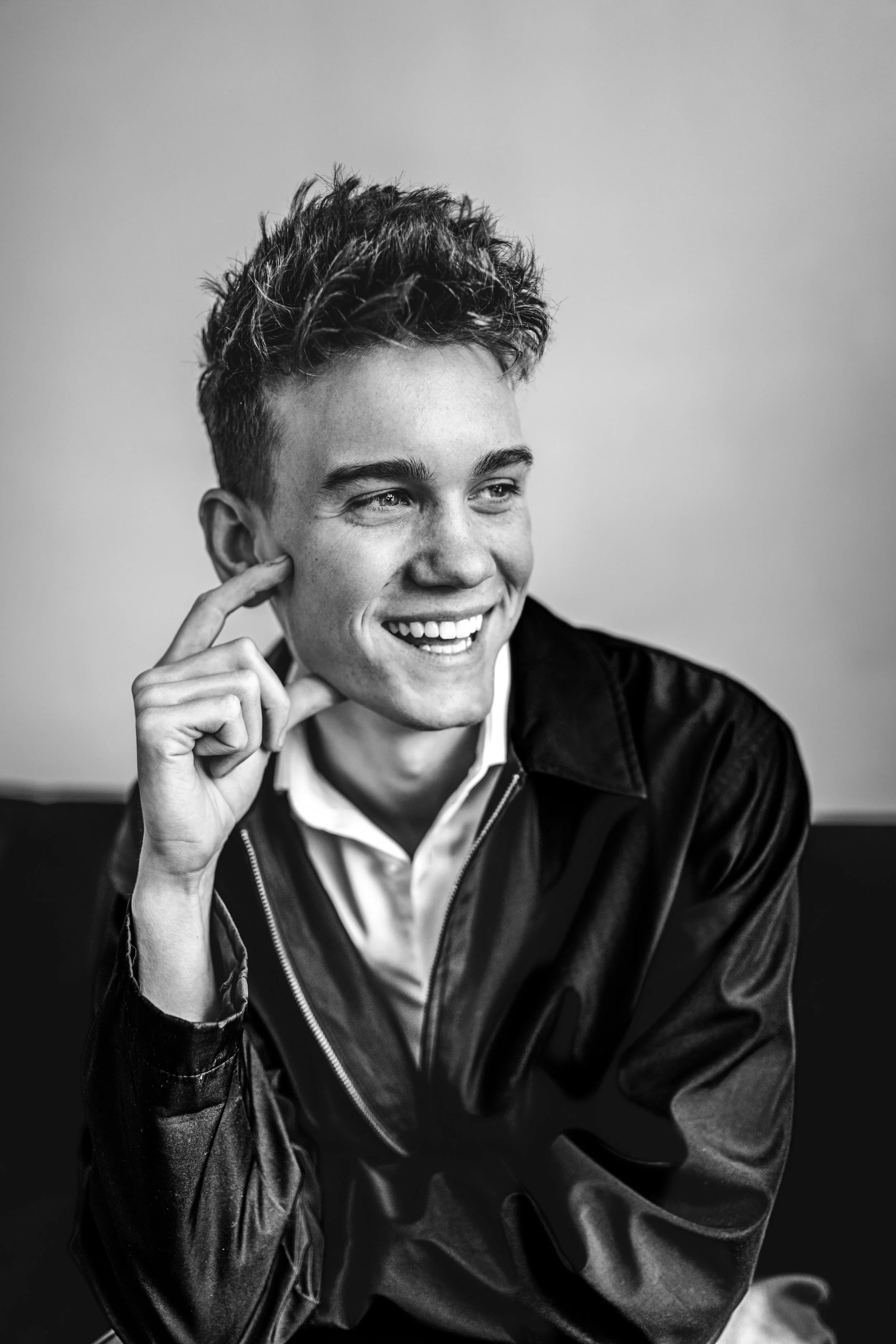 A young man smiling and touching his face with his right hand, wearing a leather jacket over a collared shirt, in black and white.
