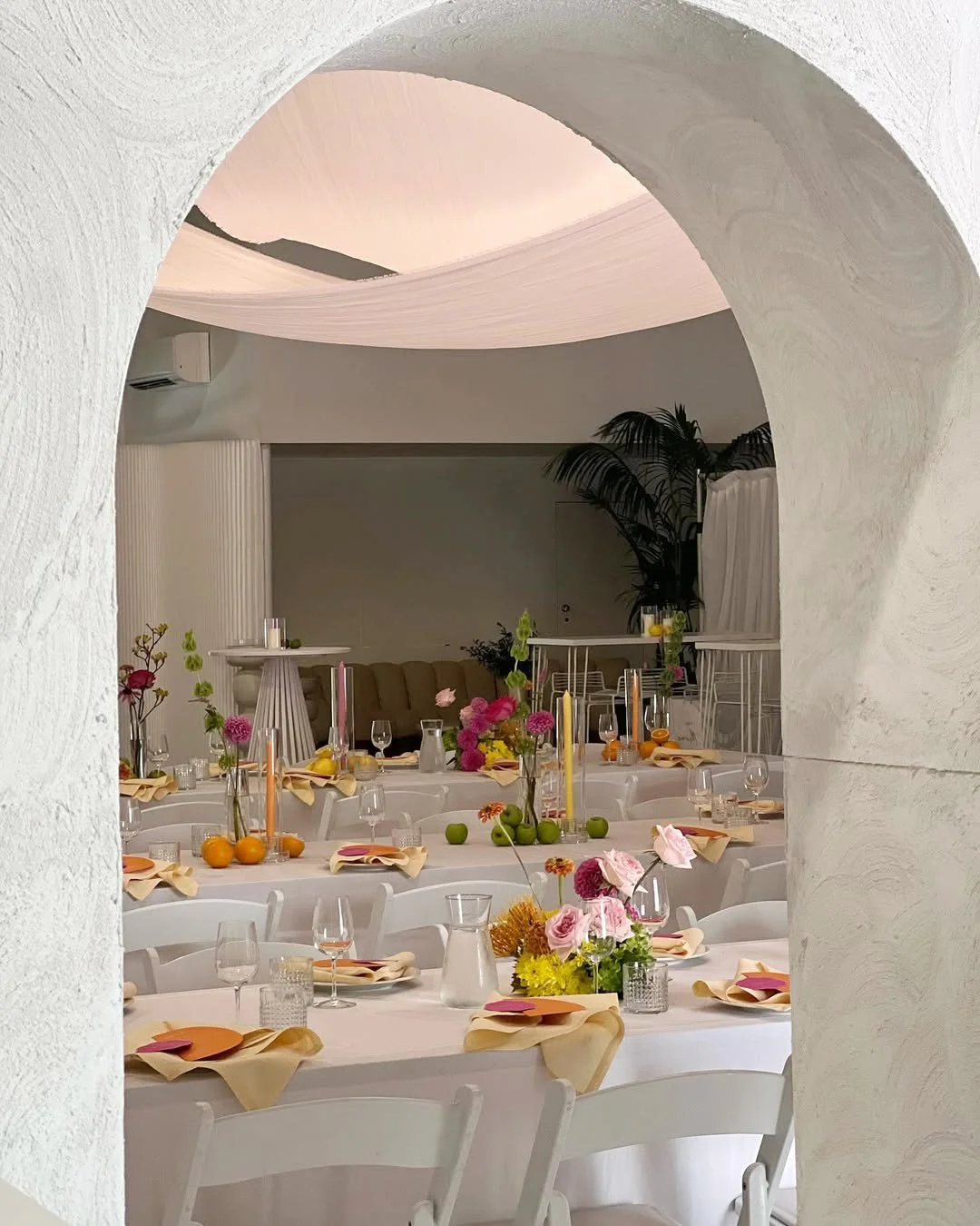 A beautifully set dining table with yellow and white flowers in vases, blue glasses, and plates with napkins, in a well-lit room with large windows.