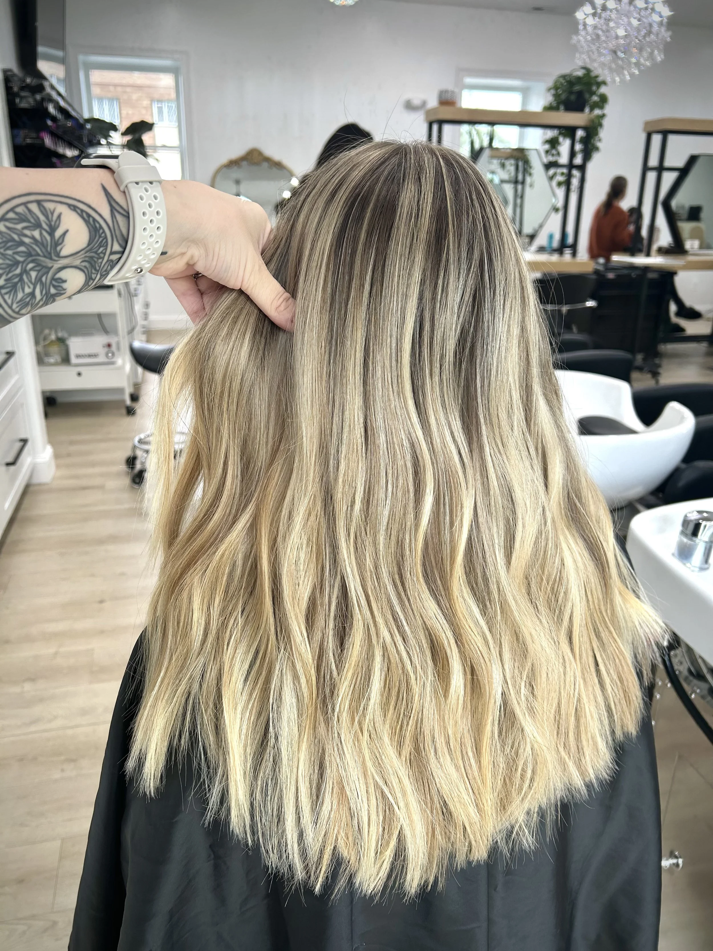 Back view of a woman with long, blonde, wavy hair in a hair salon, with a stylist's hand holding her hair. The salon has white walls, large windows, and modern decor.