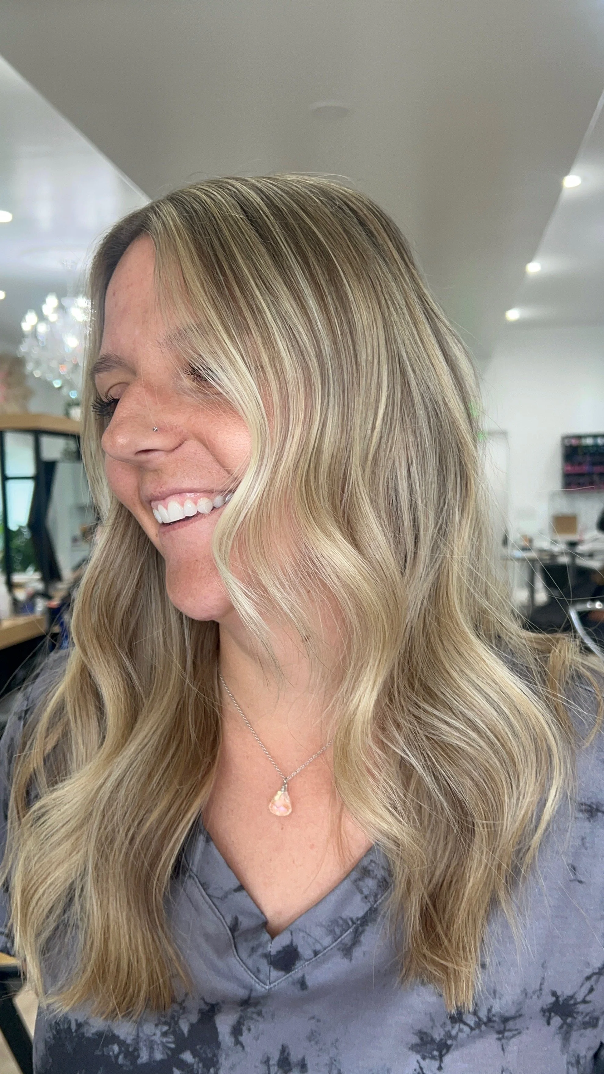 A woman with long, wavy blonde hair smiling while looking to her left, wearing a gray tie-dye shirt and a pink gemstone necklace, in a salon or studio setting.