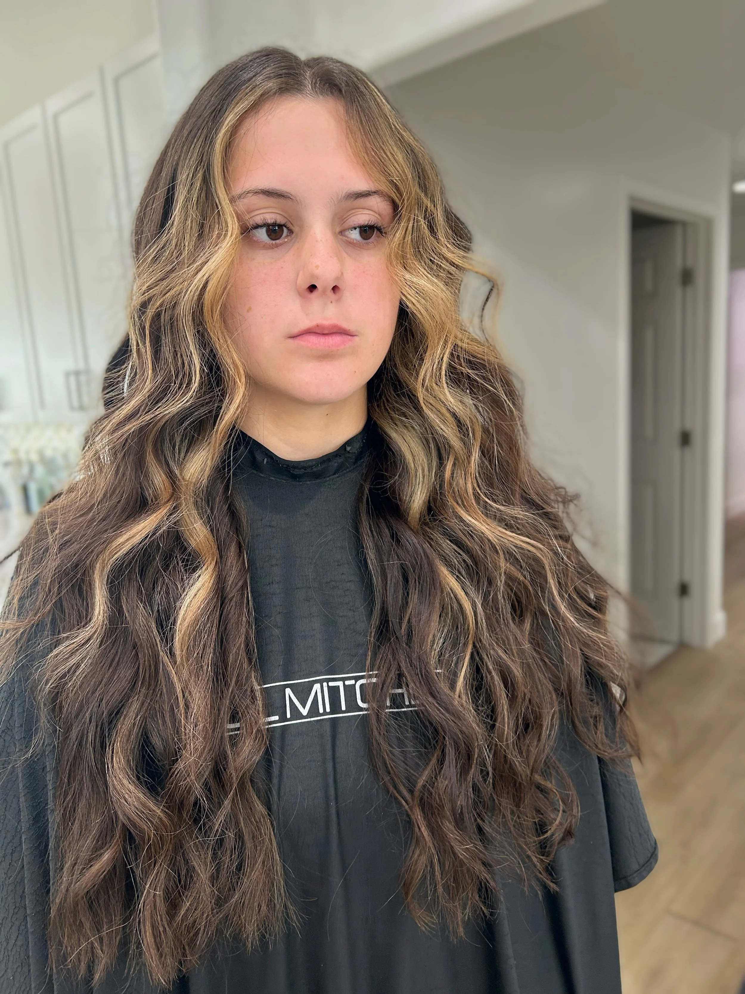 Close-up of a young woman with long, wavy brown hair with blonde highlights, wearing a black t-shirt, standing indoors near a white wall and cabinets, looking slightly to her left.