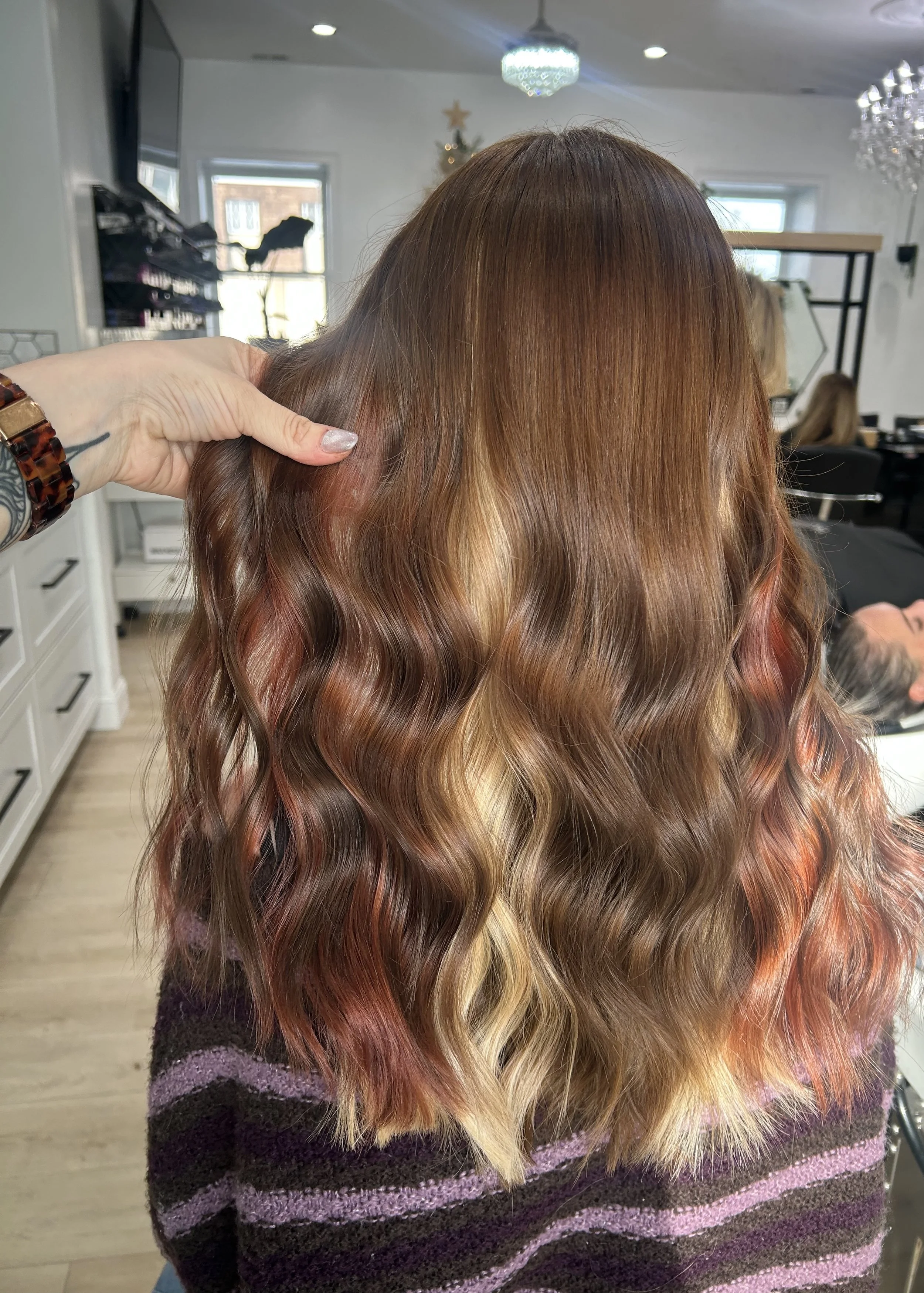 Back view of a person with wavy, multi-tonal hair in shades of brown, red, and blonde, in a hair salon.