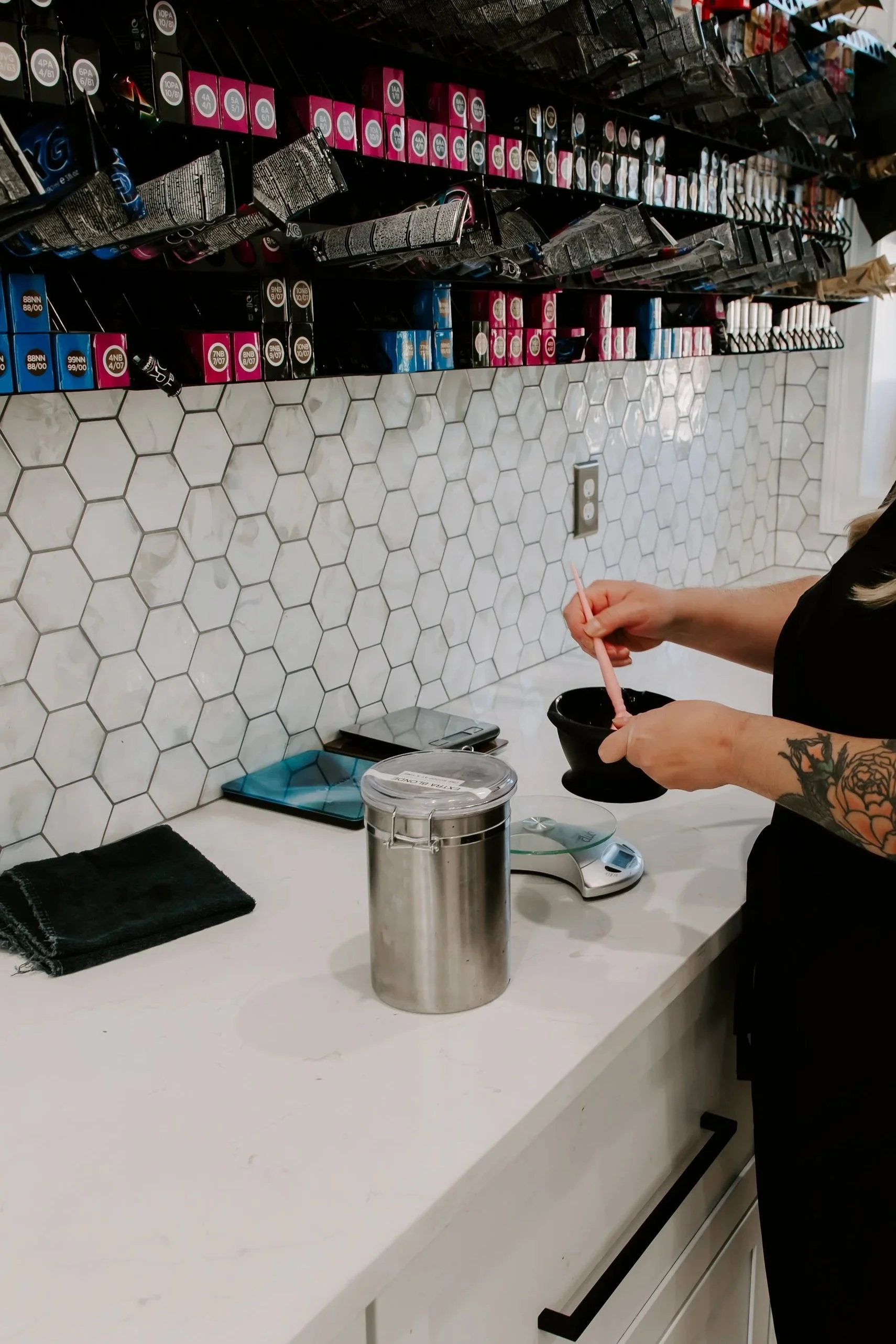 A person is preparing ingredients in a kitchen or cafe; they are holding a small black bowl and using a pink utensil. The countertop has a black towel, a digital scale, a clear cylindrical container, and a blue weighed item. The backsplash features white hexagonal tiles, and above it are shelves with various boxes and pouches, likely containing baking or cooking supplies.