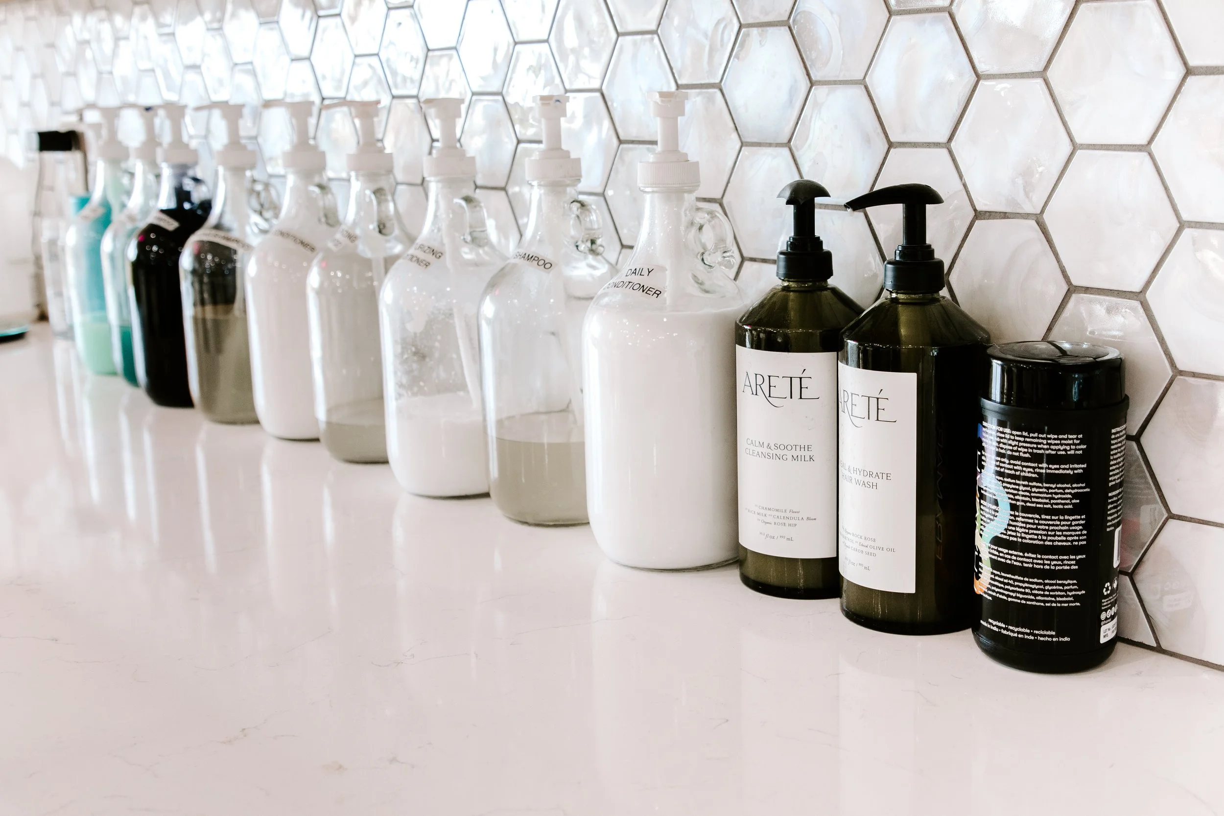 A row of soap bottles and other bathroom products on a white countertop against a hexagon-tiled wall.