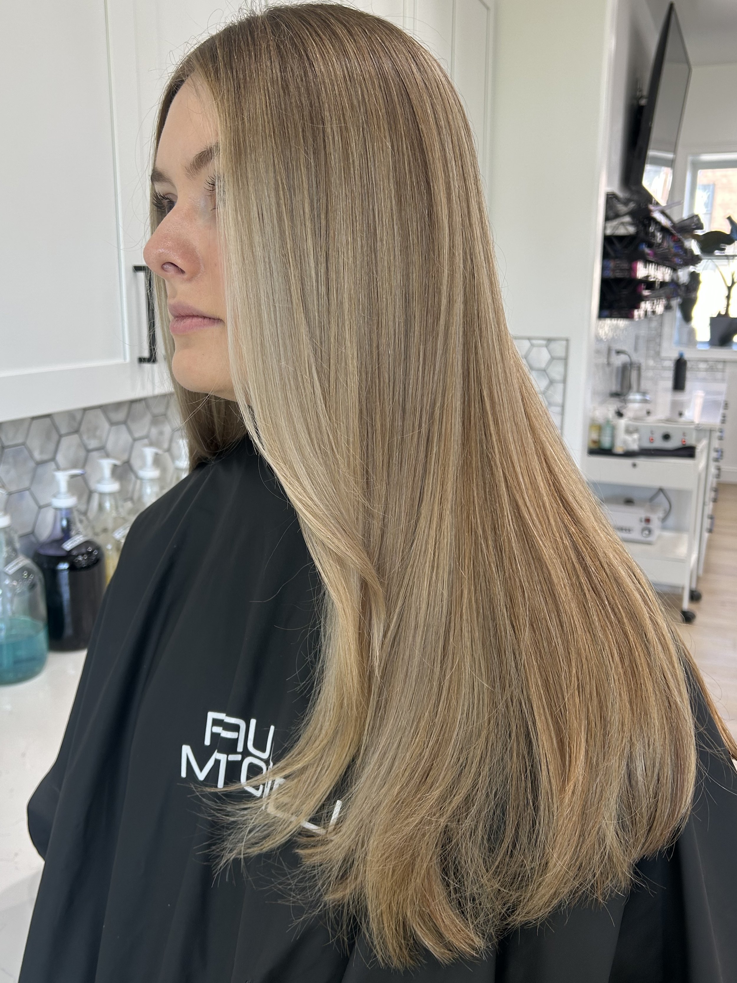 A woman with long, straight, blonde hair with darker roots. She is in a salon, wearing a black cape, with a mirror, salon supplies, and a television visible in the background.