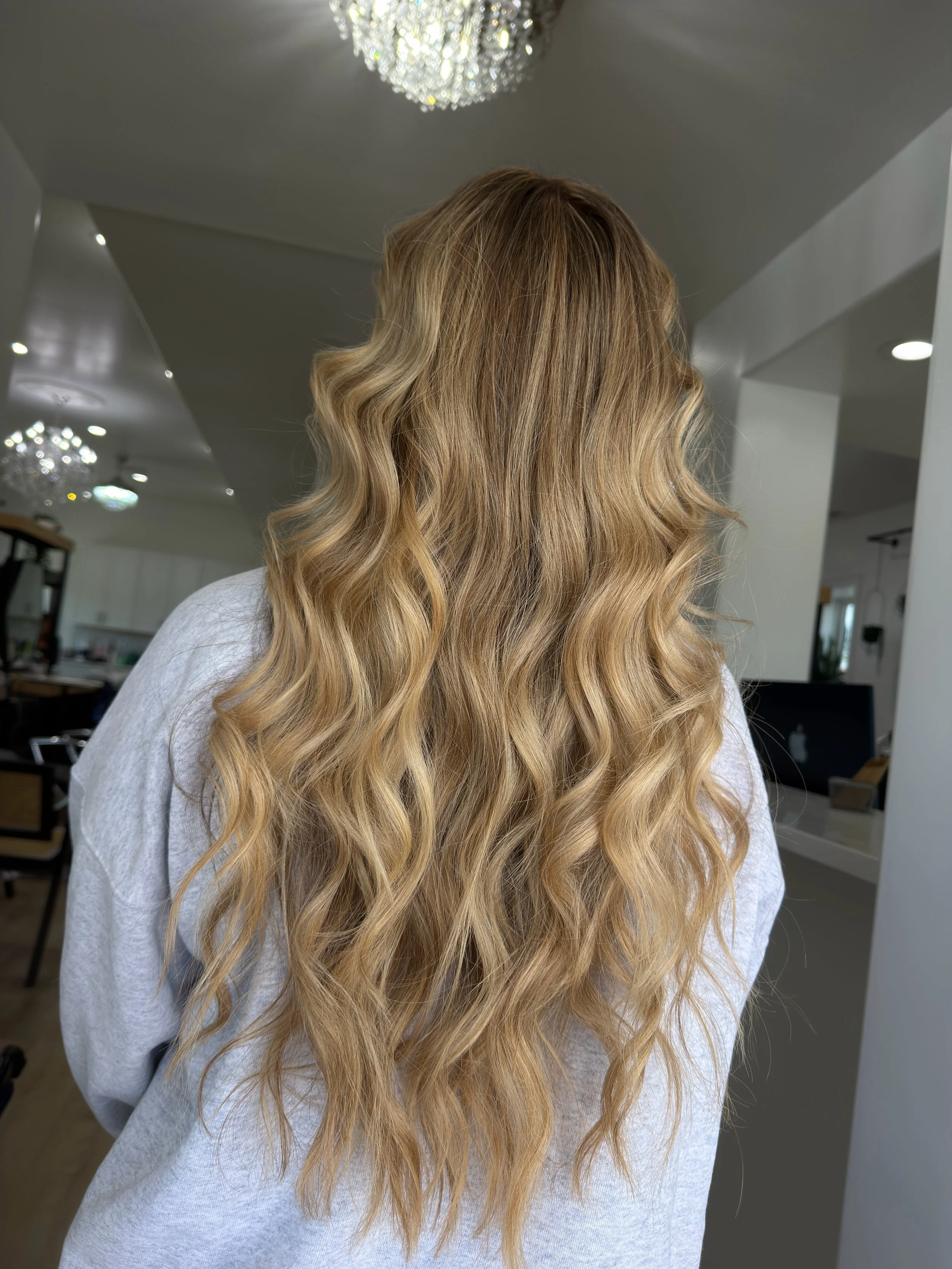 Woman with long, blonde, wavy hair standing indoors, facing away from the camera.