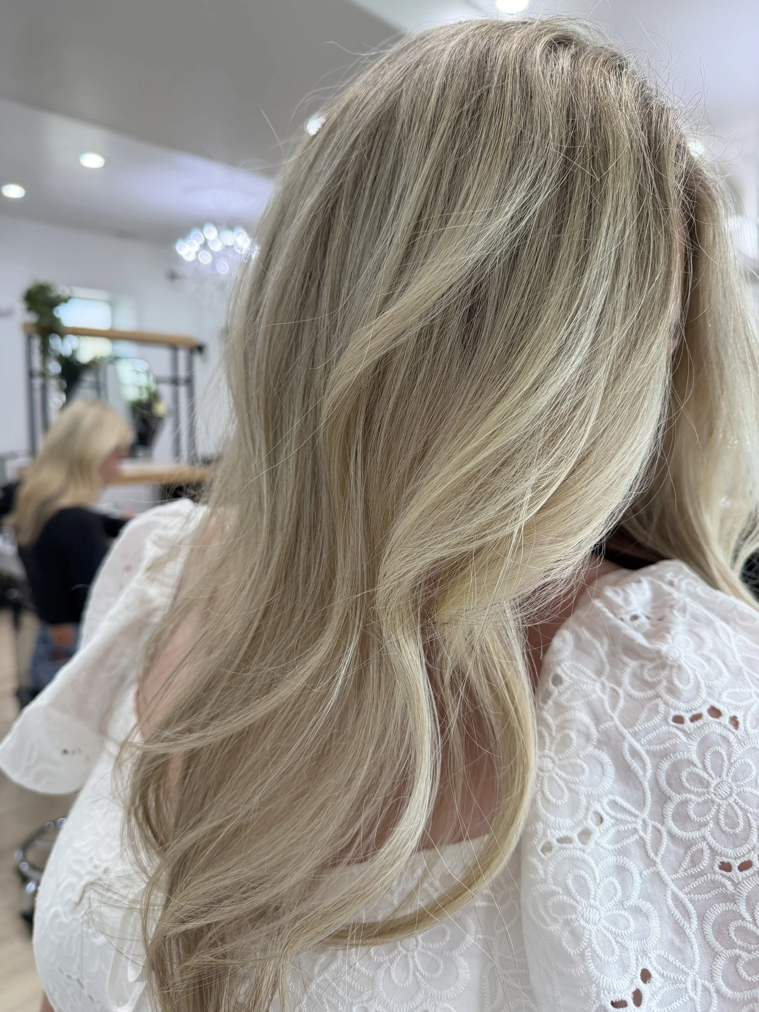 Close-up of a woman with long, wavy blonde hair wearing a white embroidered top, in a salon setting.