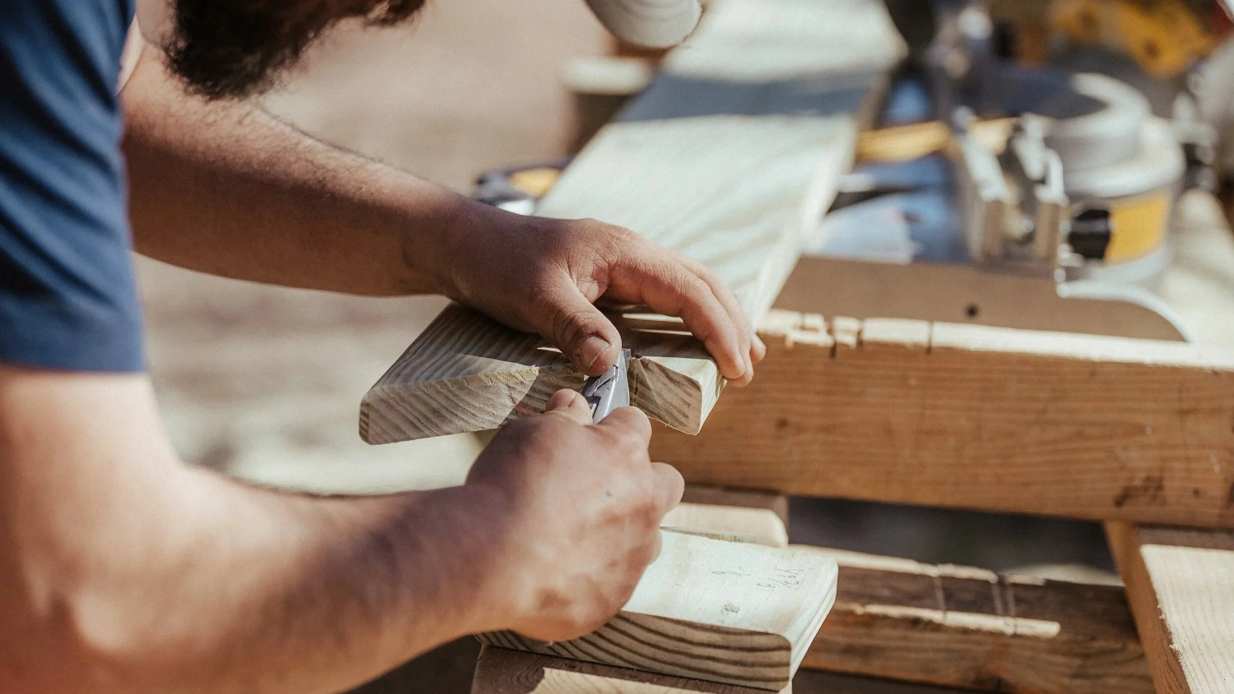 A person is woodworking, using a utility knife to carve or trim a piece of wood on a workbench, with tools and wood planks visible in the background.