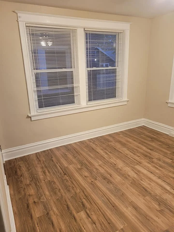 Empty room with beige walls, white trim, and wooden flooring, featuring two double-hung windows with white blinds.