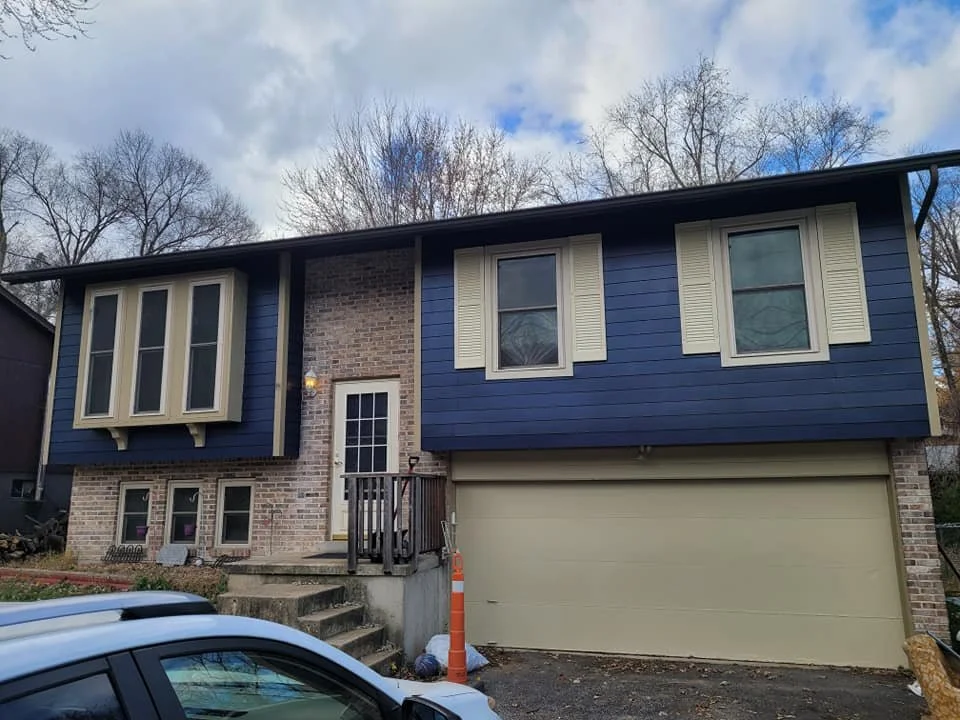 Two-story house with brick and blue siding exterior, beige garage door, and a white door with a small porch. A car is parked in front, and leafless trees are in the background under a partly cloudy sky.