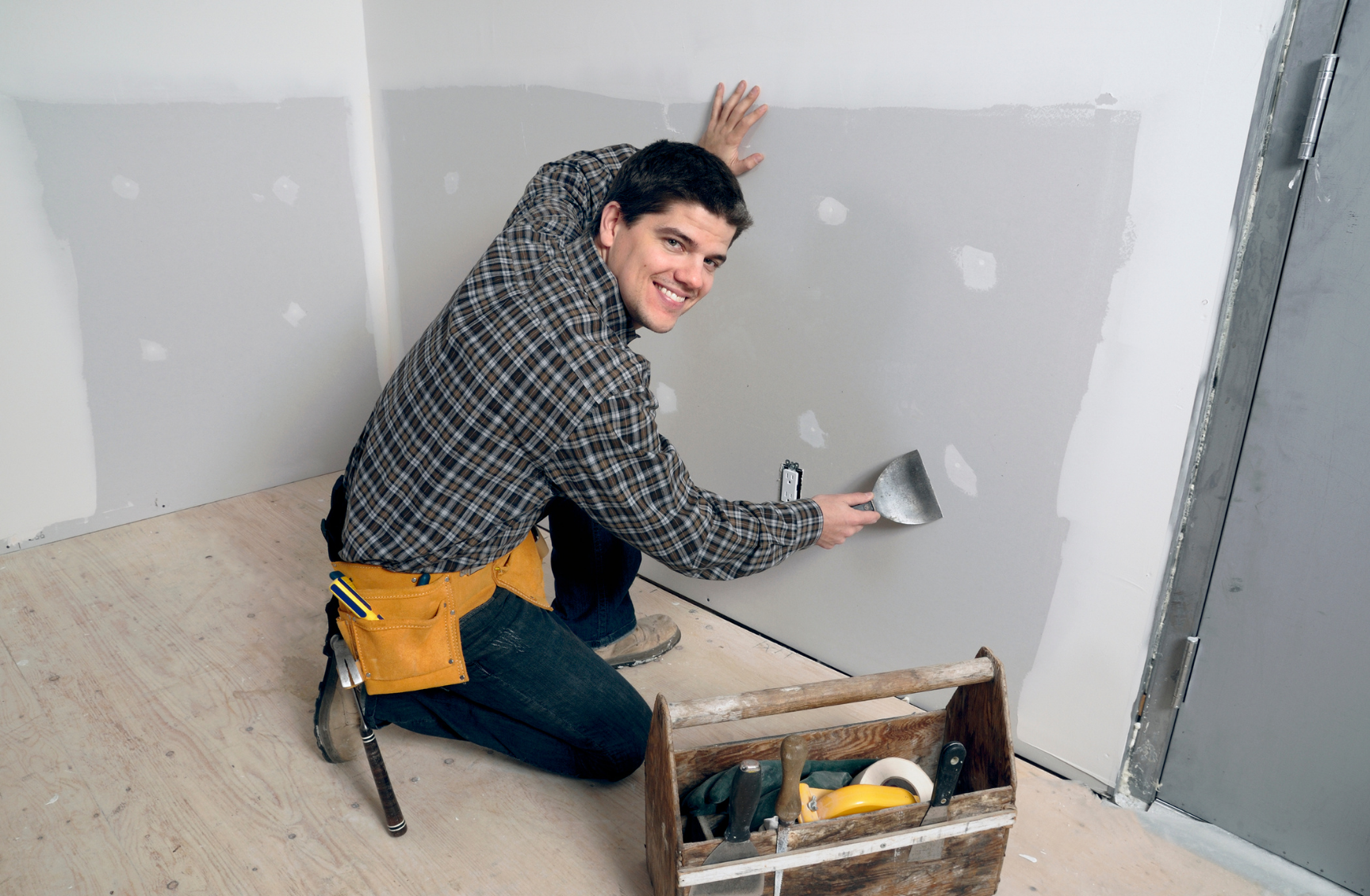 A man kneeling on a wooden floor, applying drywall compound to a wall with a putty knife, smiling at the camera, with tools in a wooden caddy nearby.