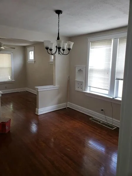 Empty living room with hardwood floors, a chandelier, multiple windows with blinds, and a small built-in shelf on the wall.
