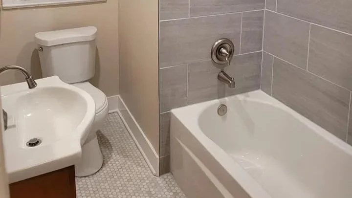 A small bathroom with a toilet, a sink, and a bathtub, with beige and light gray tiles and white fixtures.