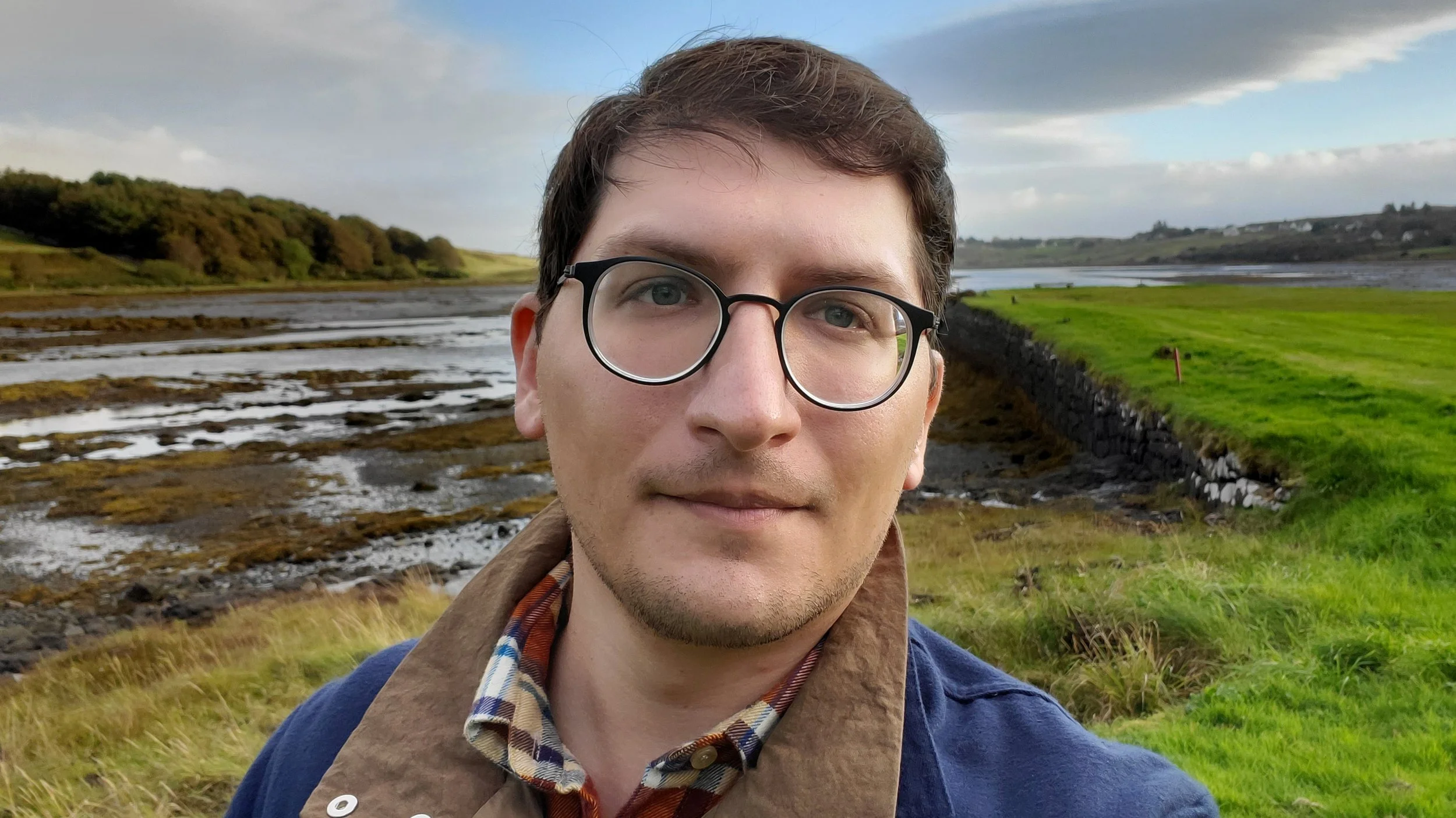 A man with glasses and a jean jacket standing in front of a green field and a cloudy blue sky