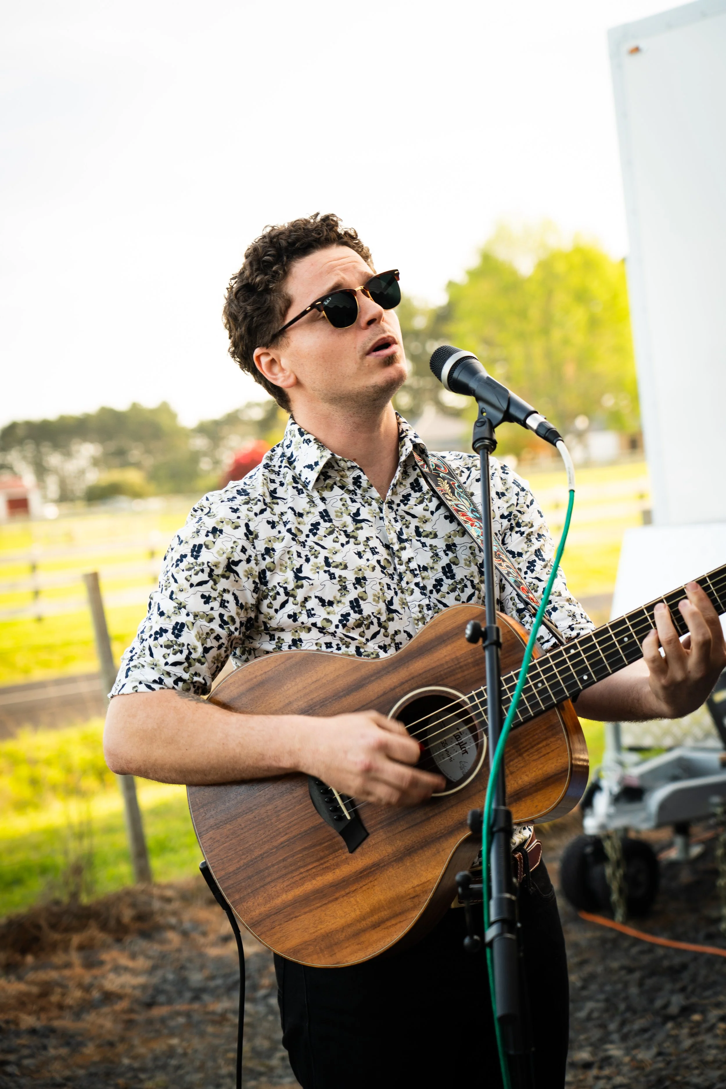 A man wearing sunglasses, a floral shirt, and playing an acoustic guitar while singing into a microphone outdoors.