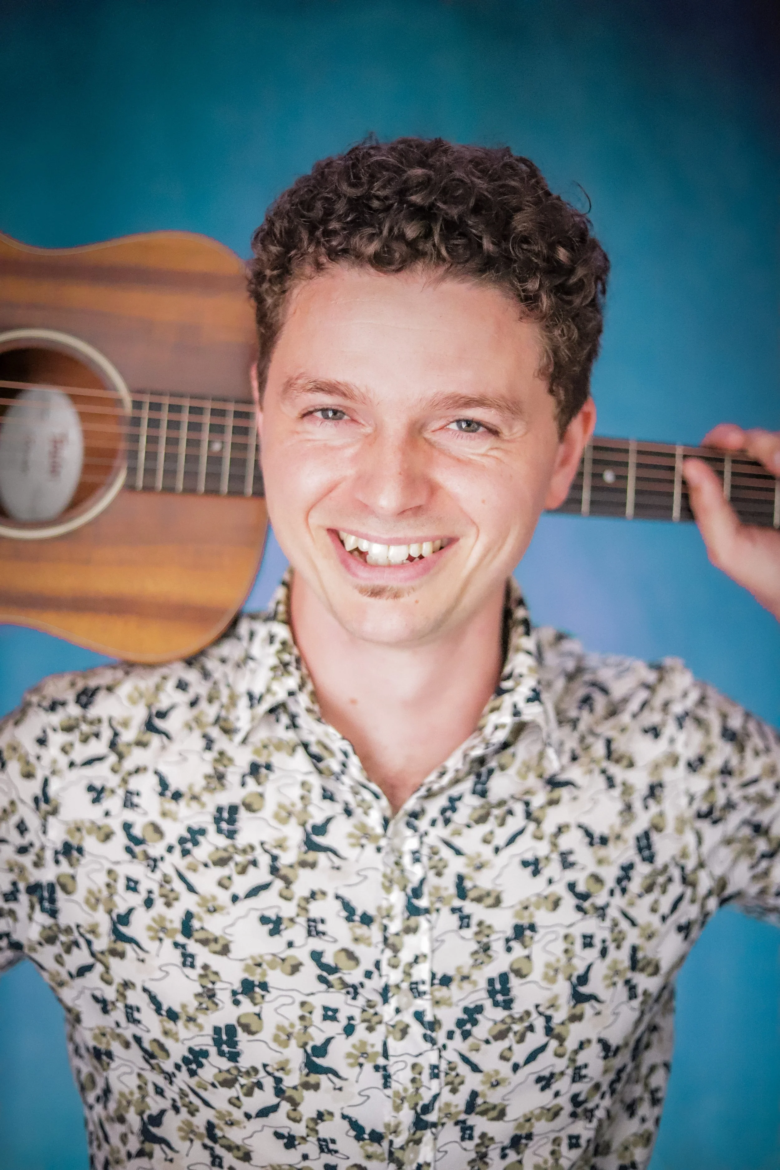 A man smiling while holding a ukulele over his shoulder, wearing a patterned shirt, against a blue background.