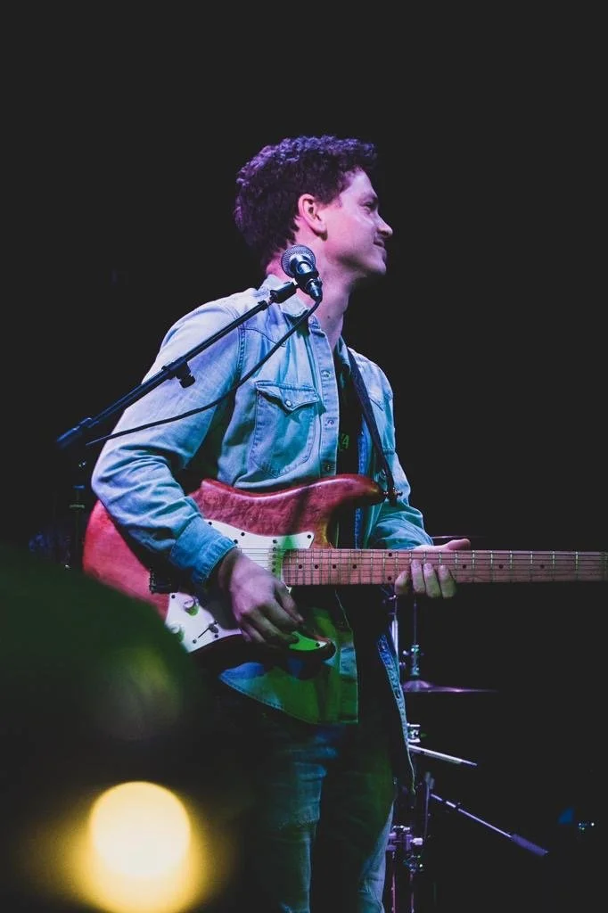 A person playing an electric guitar on stage with a microphone nearby, wearing a denim jacket and dark pants, against a dark background.