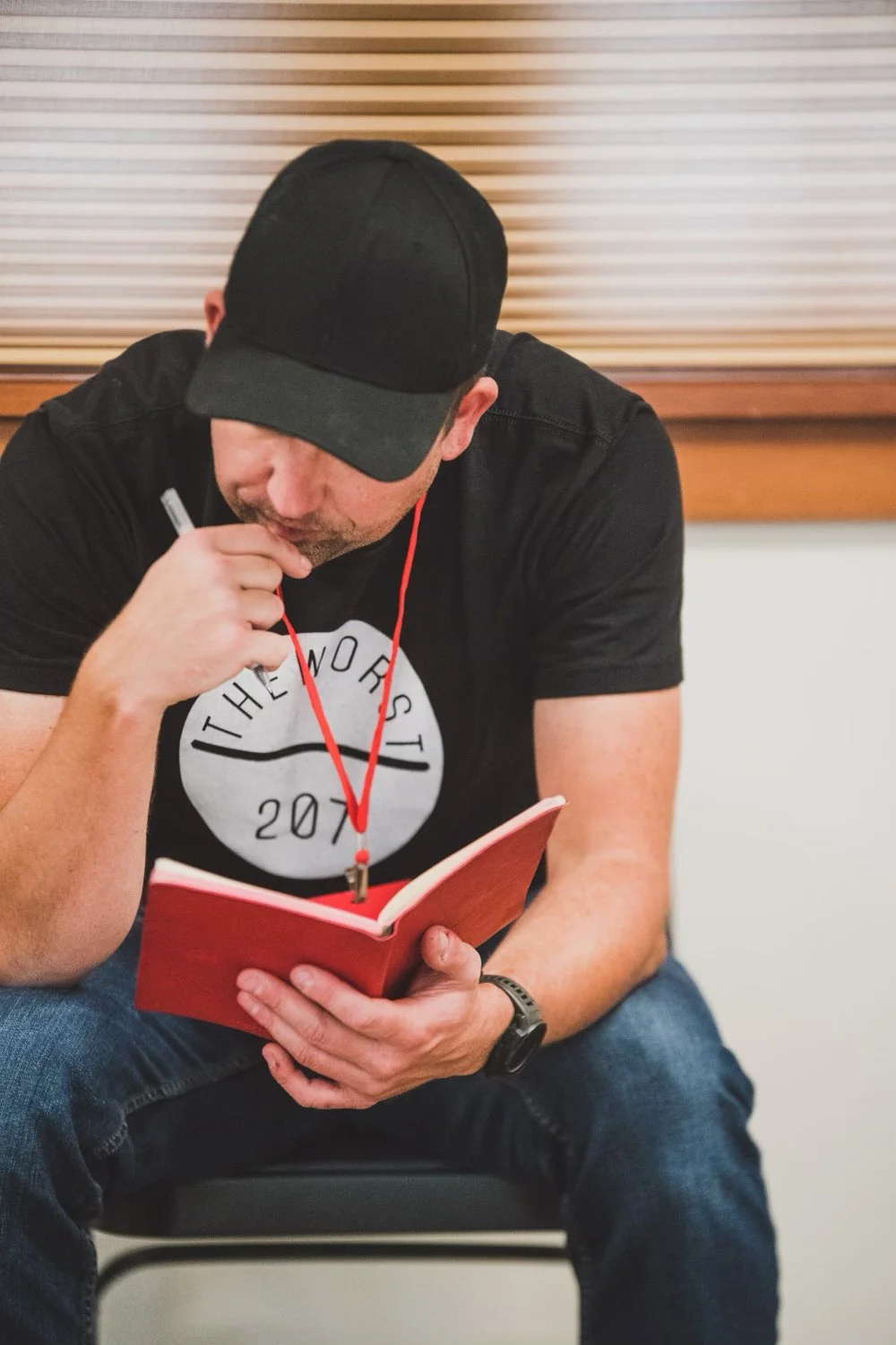 Ben Miosi sitting on a chair, reading writing in his red design journal, wearing a black cap, black t-shirt, and jeans, with a red Bend Design Week lanyard around his neck, in a room with beige walls and closed horizontal blinds.