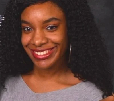A woman with dark, curly hair smiling and wearing a gray top.