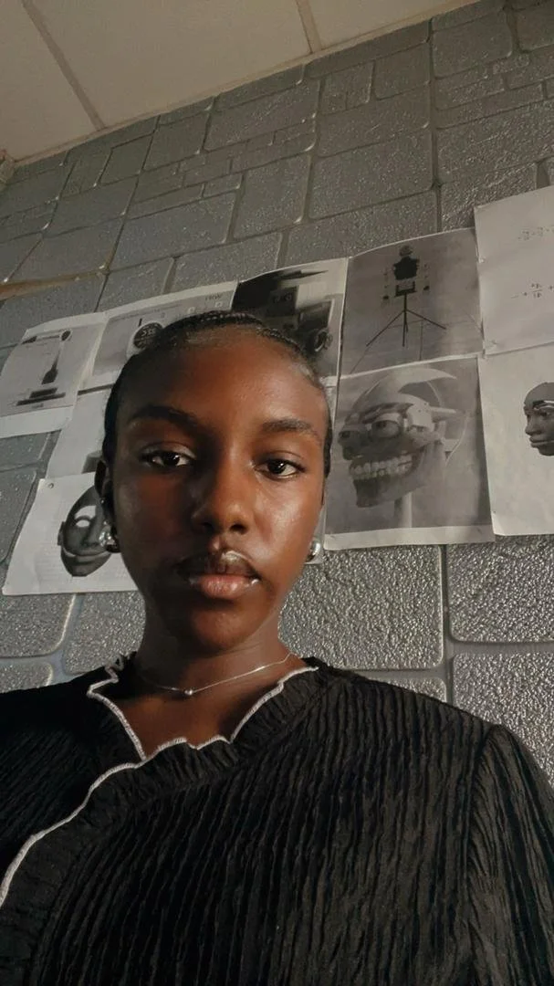 A young woman with braided hair in a bun, wearing a black textured top with white trim and a necklace, stands in front of a wall with anatomical sketches of skulls and faces on gray cinder block.