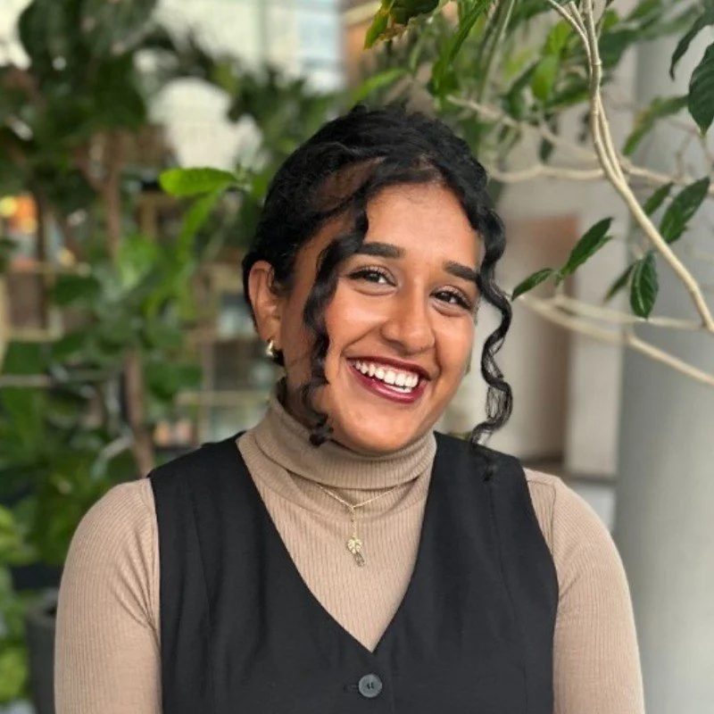 Smiling person with curly hair, wearing a beige turtleneck and black vest, standing in front of greenery.