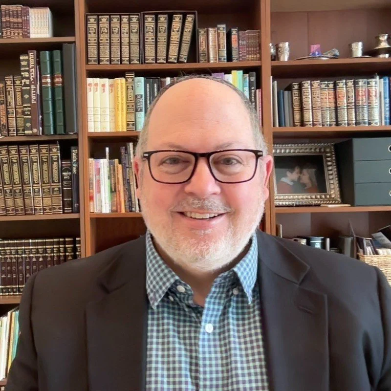 A middle-aged man with glasses, a beard, and a bald head smiling at the camera in front of a bookshelf filled with books and decorative items.