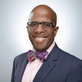 Professional headshot of a smiling man in a suit and bowtie, wearing glasses.