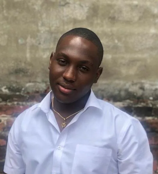 Young man with short hair wearing a white shirt and a gold necklace, standing outdoors in front of a weathered concrete wall.