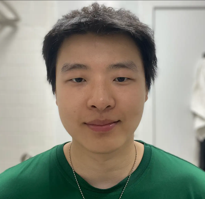 A young man with short black hair, wearing a green shirt and a silver chain, smiling at the camera in an indoor setting.