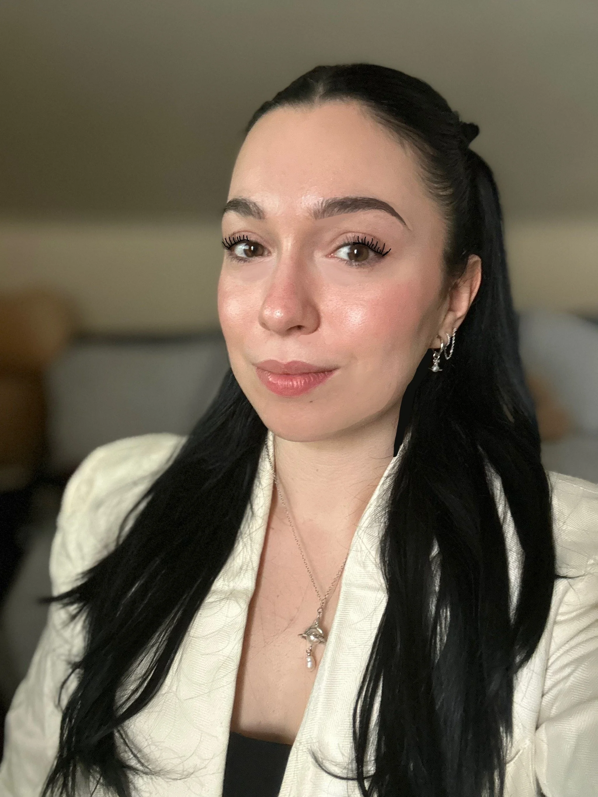 Close-up of a woman with long black hair, wearing a cream-colored jacket, silver earrings, a necklace with a dolphin pendant, and makeup with defined eyebrows and eyelashes.
