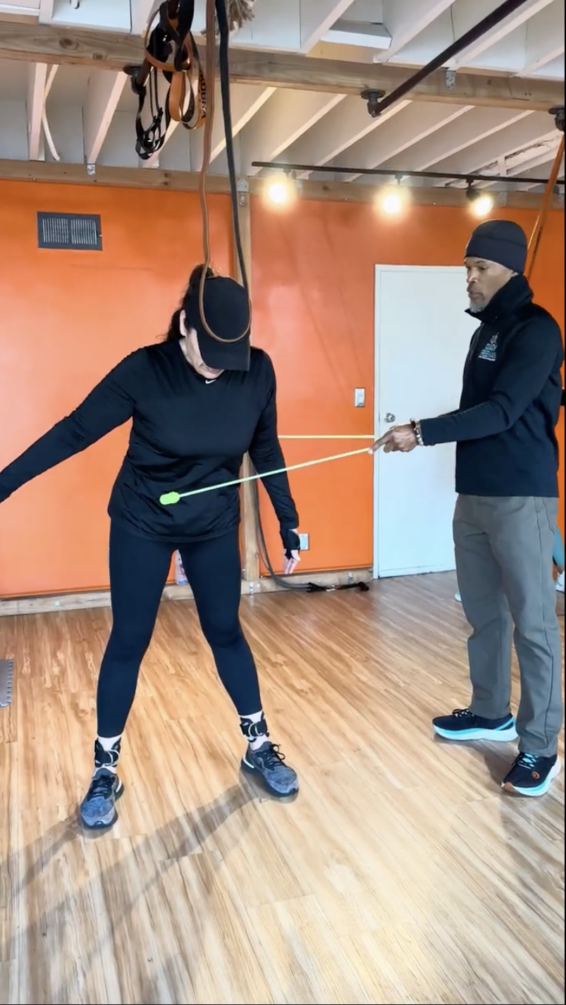 A woman is participating in a physical therapy session, using a green aquatic paddle and wearing a black cap, black long-sleeve shirt, black leggings, and athletic shoes. A man, likely a therapist, stands beside her, holding the paddle and observing her. The room has an orange wall, a wooden floor, and therapy equipment hanging from the ceiling.