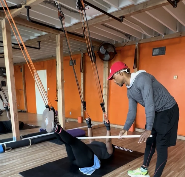 Personal trainer assisting a woman with resistance band exercises in a gym studio with orange walls and wooden beams.