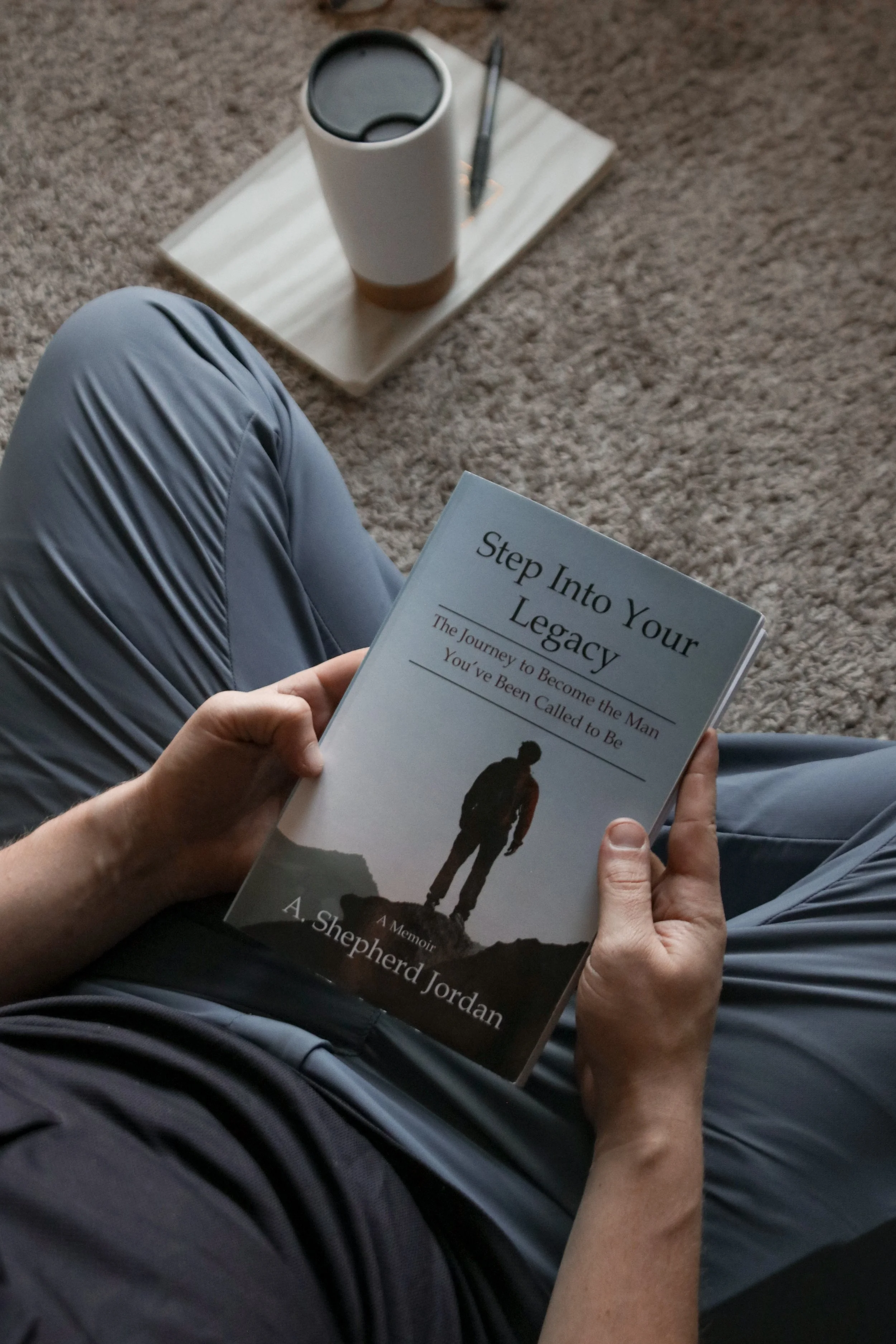 Person sitting cross-legged on a carpeted floor, holding a book titled 'Step Into Your Legacy' by A. Shepherd Jordan. Next to them is a coffee cup, a notebook, and a pen on a wooden tray.