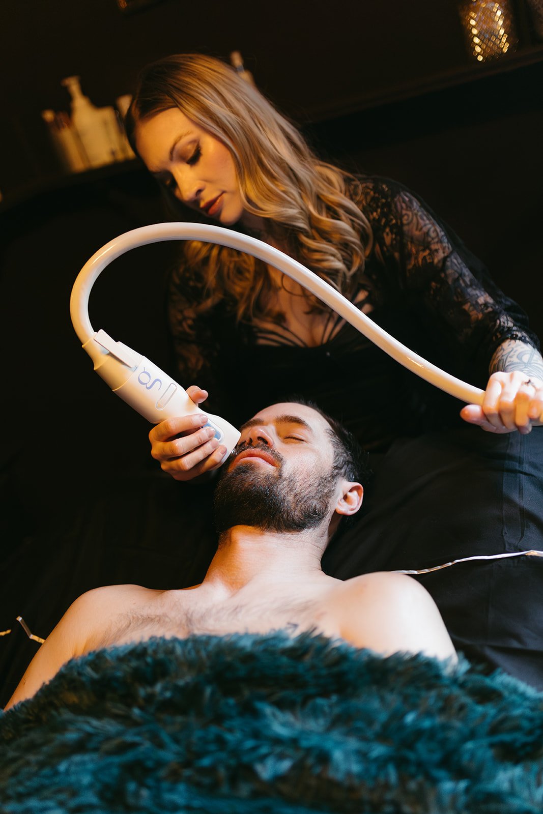 A woman with long, wavy hair and tattoos on her arm is using a medical device on a man with a beard who is lying down with his eyes closed.