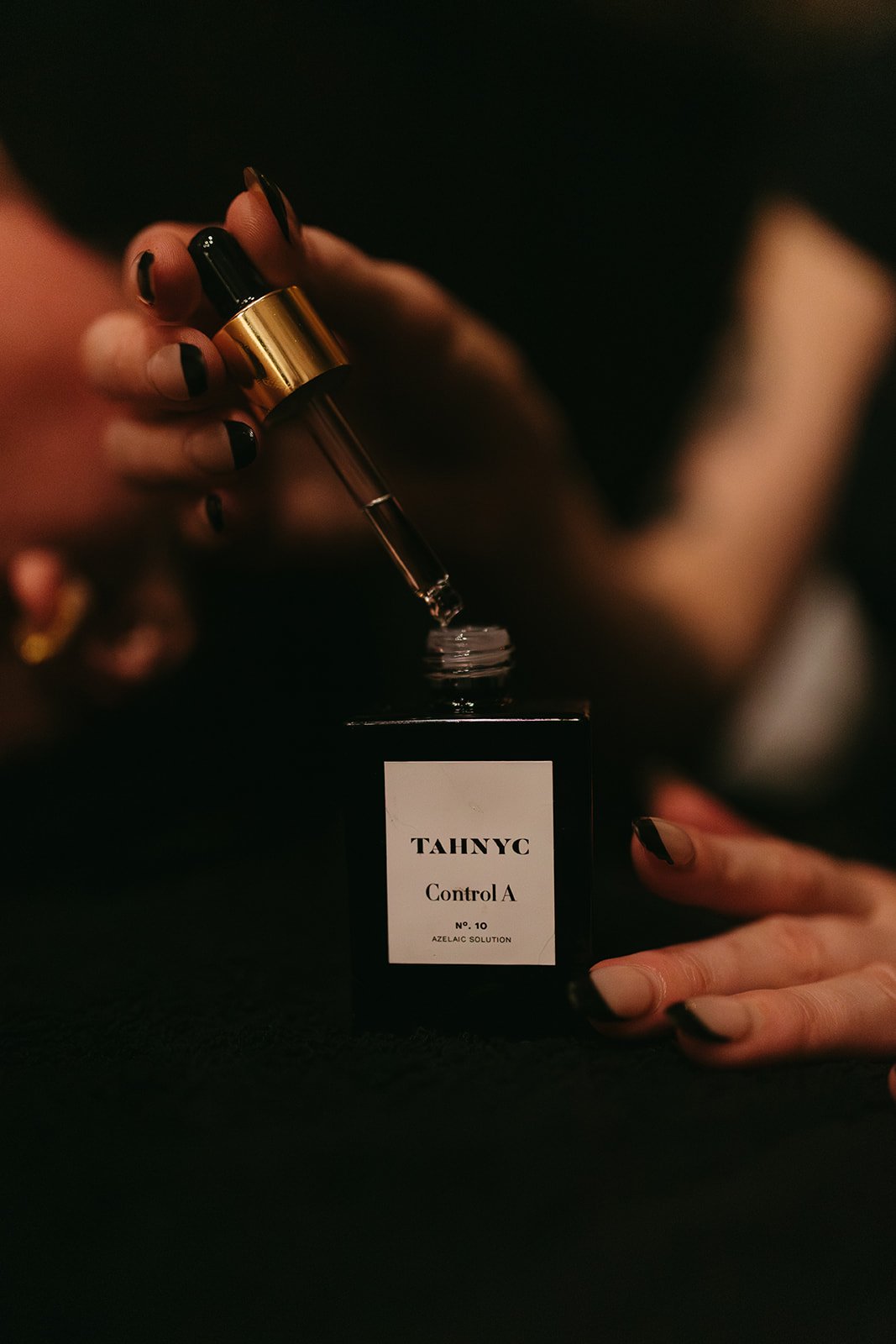 Person with black and beige painted nails using a dropper to dispense liquid from a bottle labeled 'TAHNYC Control A No. 10 Azelaic Solution'.