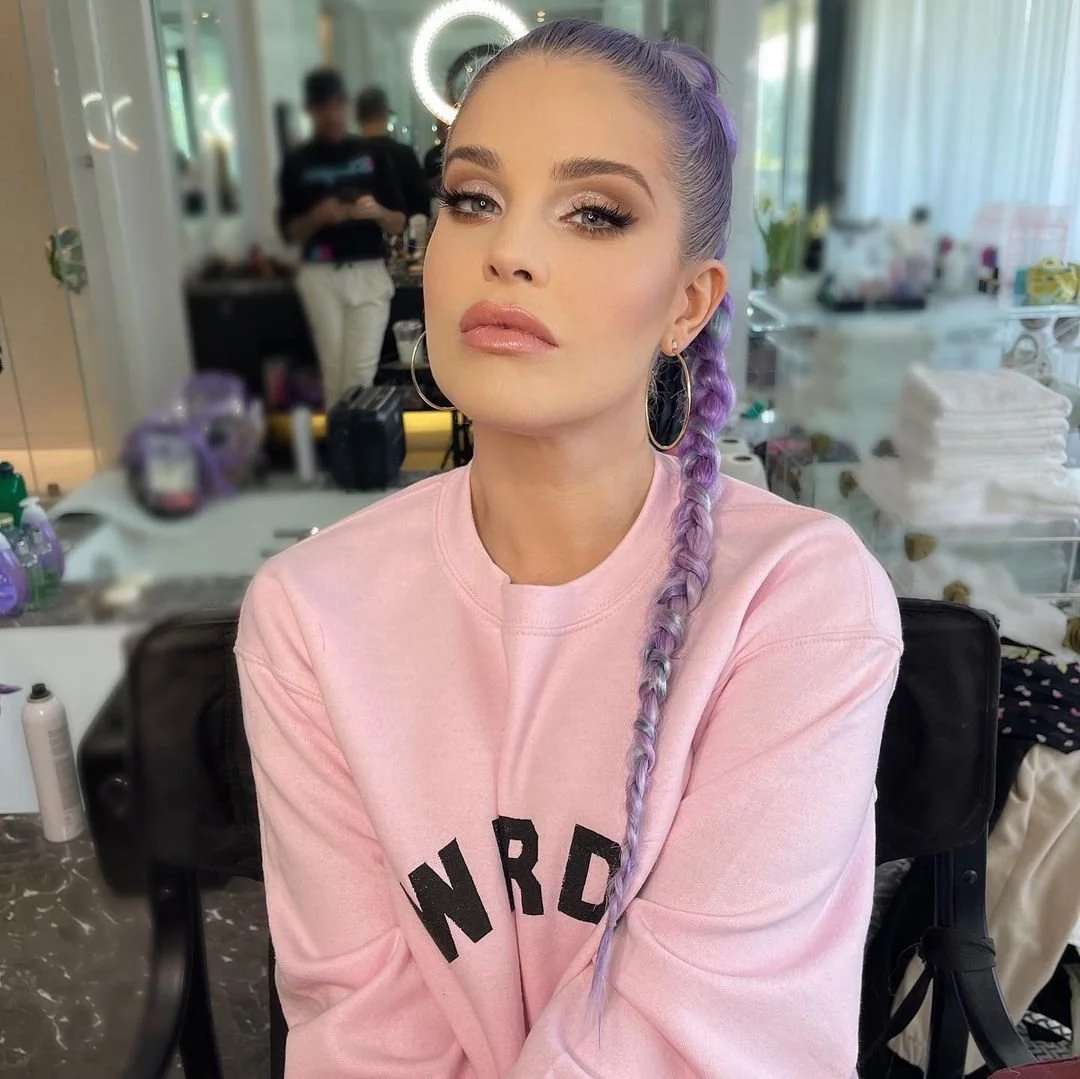 A person with purple braided hair wearing a pink sweatshirt, sitting in a salon or makeup studio. Reflection of a person in the background. The room has styling tools and towels.