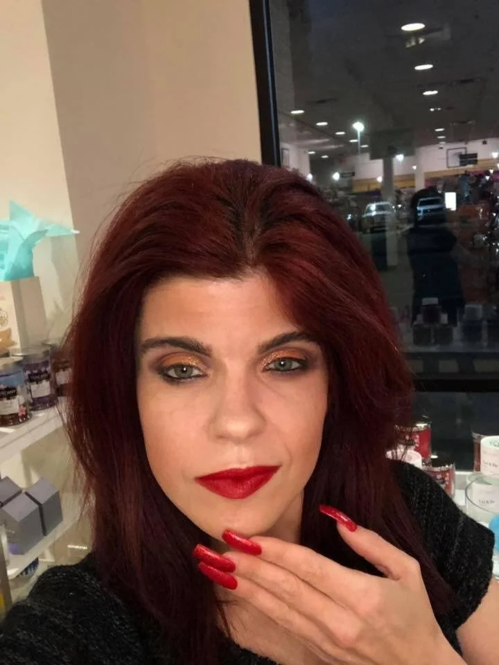 Person with long red nails, red lipstick, and dark eye makeup, inside a room with shelves in the background.