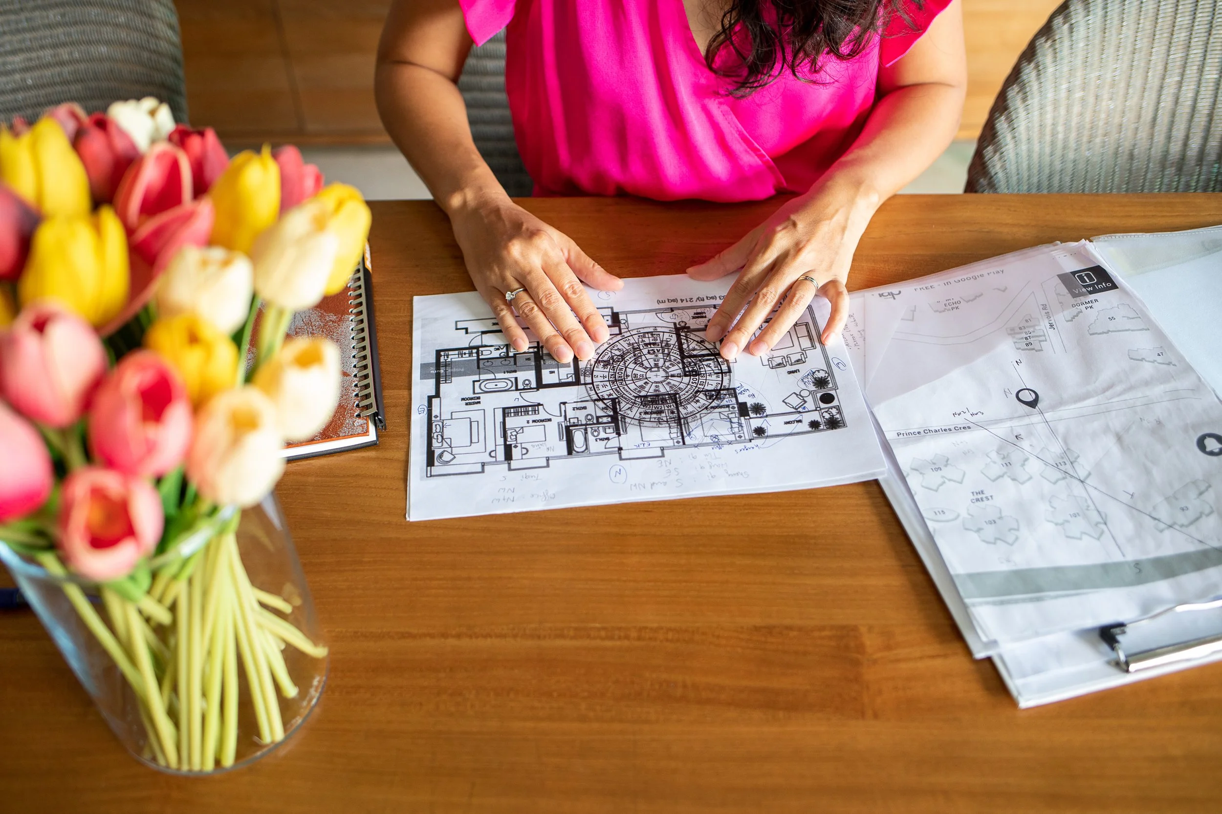 A person in a pink shirt working on building plans at a wooden table. A bouquet of pink and yellow tulips is in the foreground.